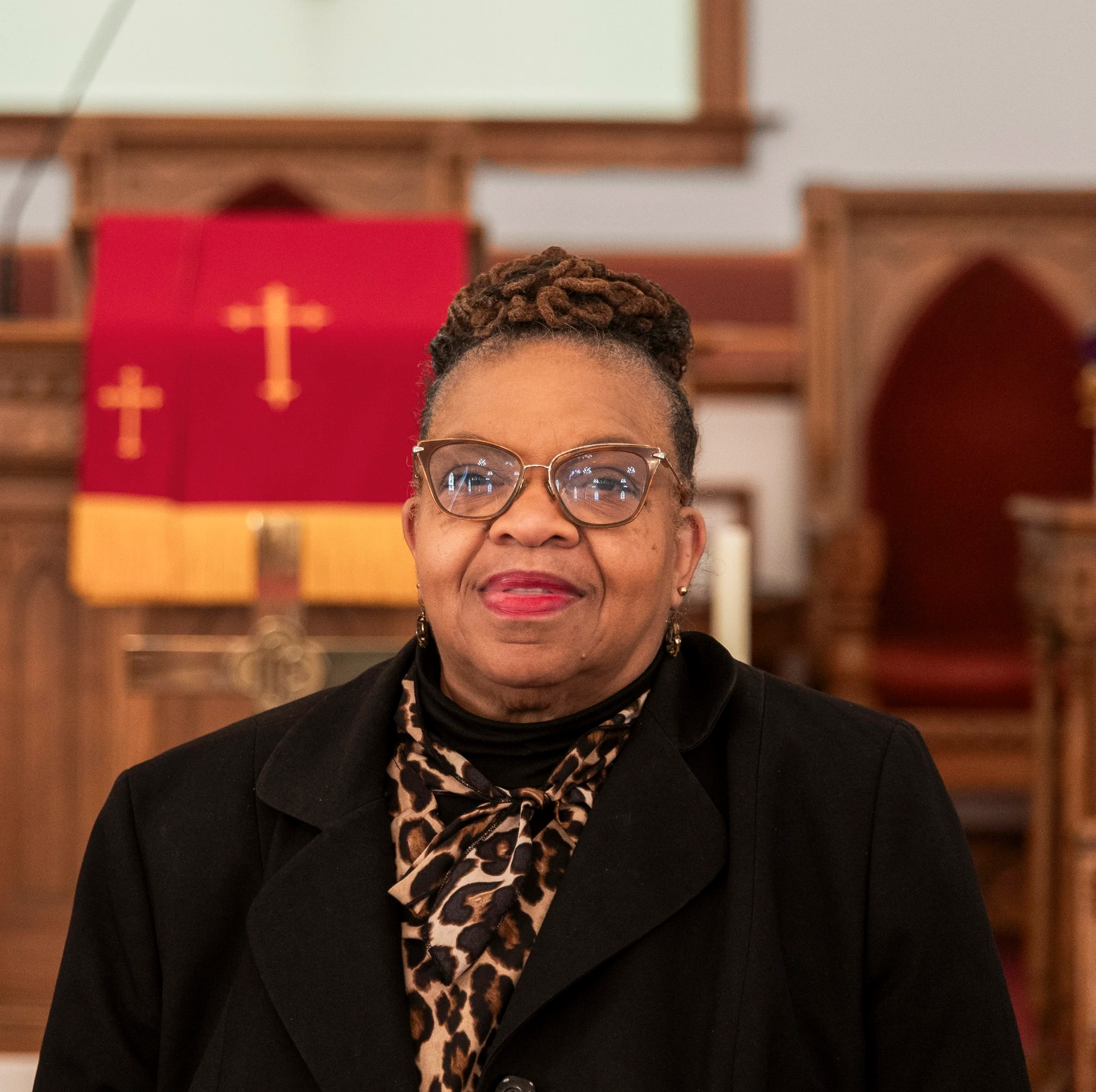 A woman with glasses and earrings, wearing a black blazer and a leopard print scarf, smiling inside a church with wooden pews and red banner with crosses in the background.