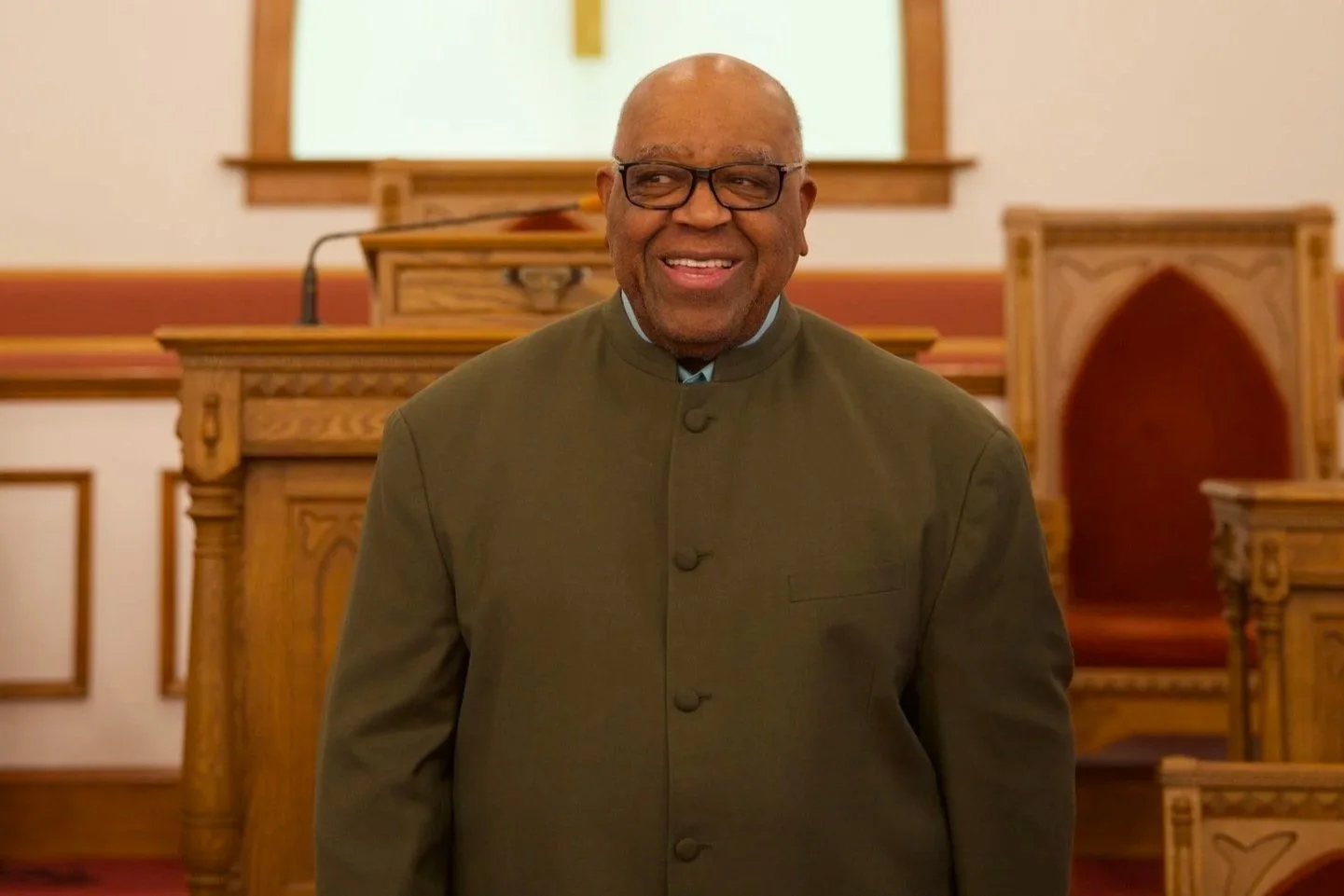 An elderly man wearing glasses and a traditional green Chinese suit stands in a church, smiling. The church has wooden pews and spiritual artwork.