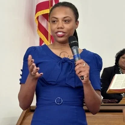 A woman speaking into a microphone, wearing a blue dress, with the American flag in the background and a woman with a book in the background.