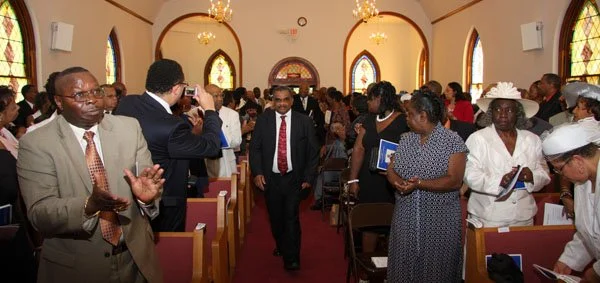 People gathered in a church, some standing and some seated, with a man in a red tie walking down the aisle.