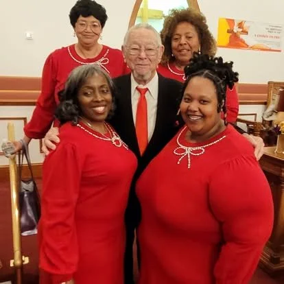 Five people, four women wearing red dresses and one elderly man in a black suit with a red tie, pose together indoors, smiling for the camera.