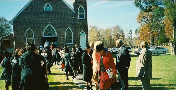 People standing in line outside a church on a sunny day, with trees and parked cars in the background.