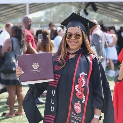 Smiling young woman in graduation cap and gown holding diploma with a crowd of people in the background.