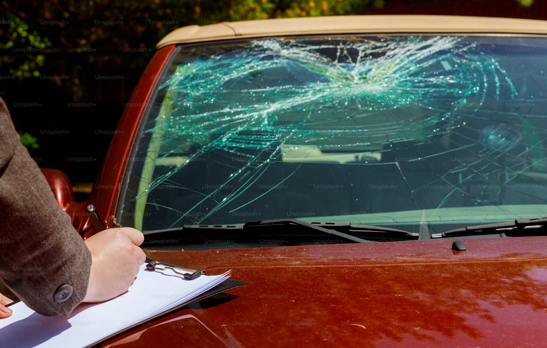 A person holding a clipboard and pen while inspecting a heavily cracked and shattered windshield of a red car, with a police or investigator present.