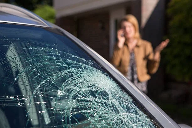 Close-up of a car with a shattered windshield, with a woman in the background talking on a cellphone, raising her hand.