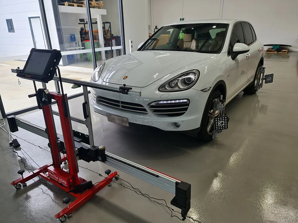 A white Porsche Macan SUV in a testing facility with wheel alignment equipment attached to its wheels.