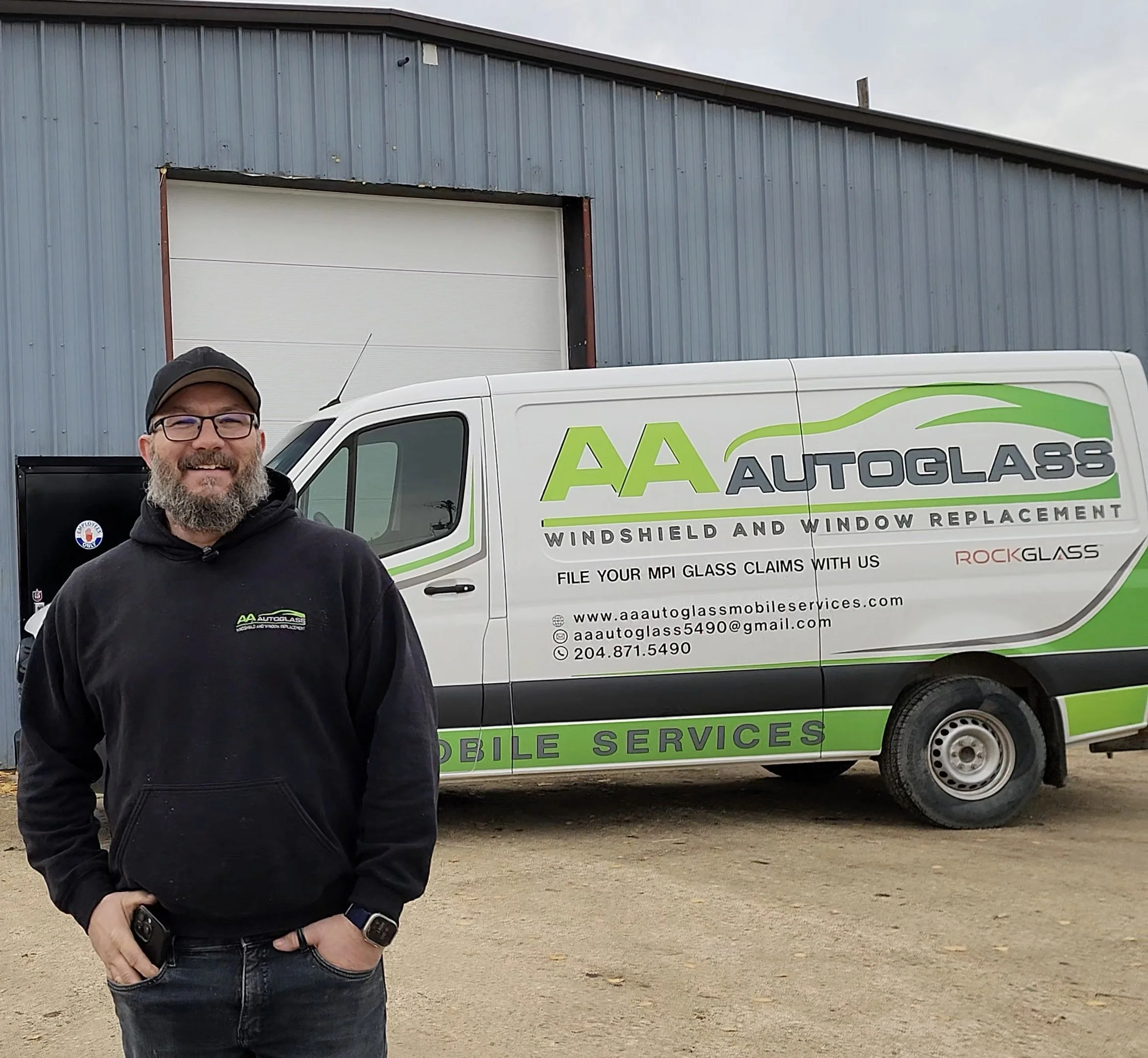 A smiling man with glasses and a beard standing in front of a white AA Autoglass service van with green and black branding, outside a blue industrial building.