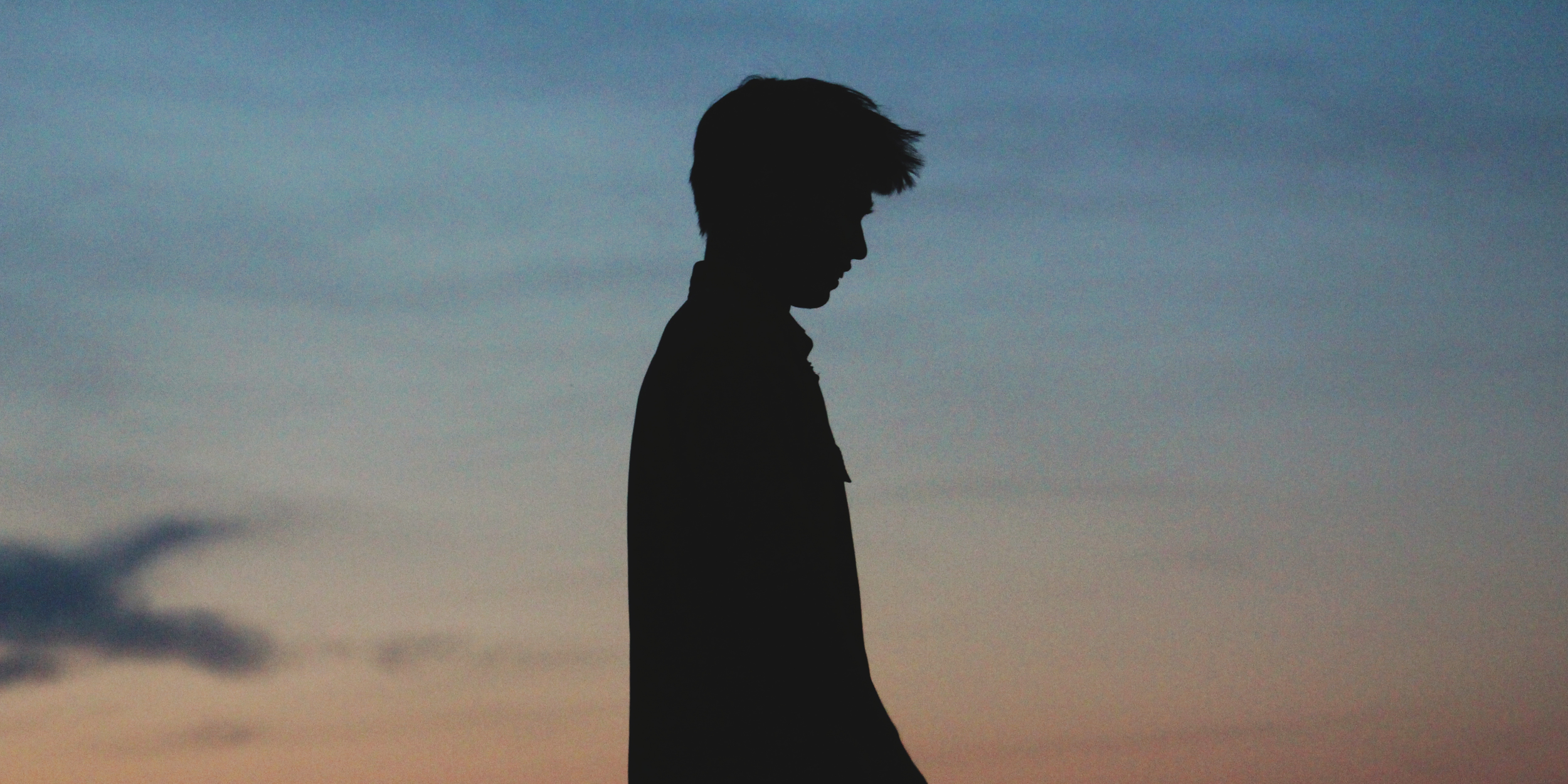 Silhouette of a young man with a textured hairstyle looking down against a sunset sky with clouds.