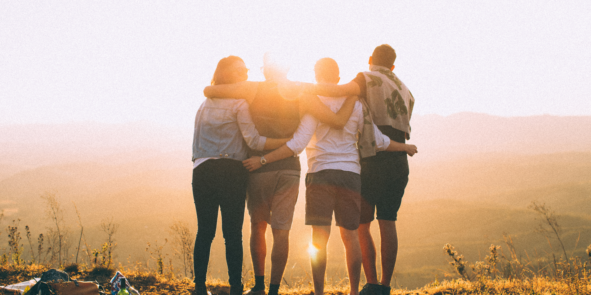 A group of five young friends standing together on a hill at sunset, with their arms around each other, enjoying the view and each other's company.