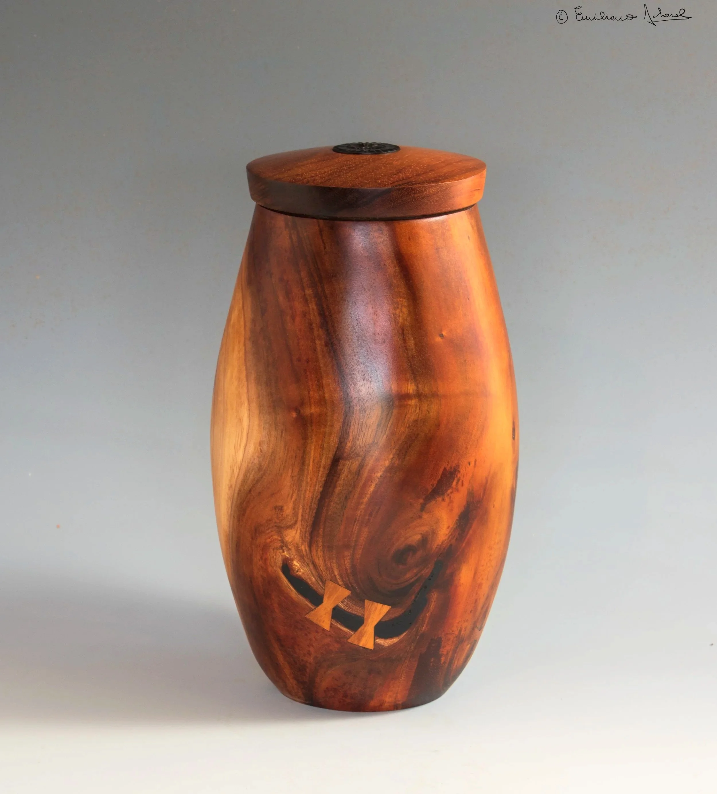 native Hawaiian Koa wood Urn. vase with a rounded, slightly tapered shape, featuring a carved face with a wide, smiling mouth and closed eyes, against a plain gradient background.