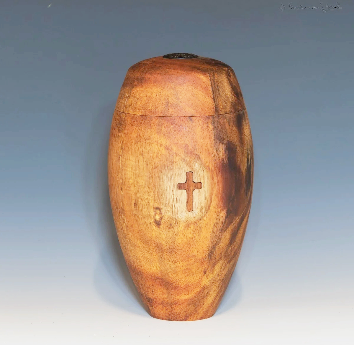A native Hawaiian Koa tree wood urn with a carved cross on the front, smooth and polished surface, against a gradient blue background.