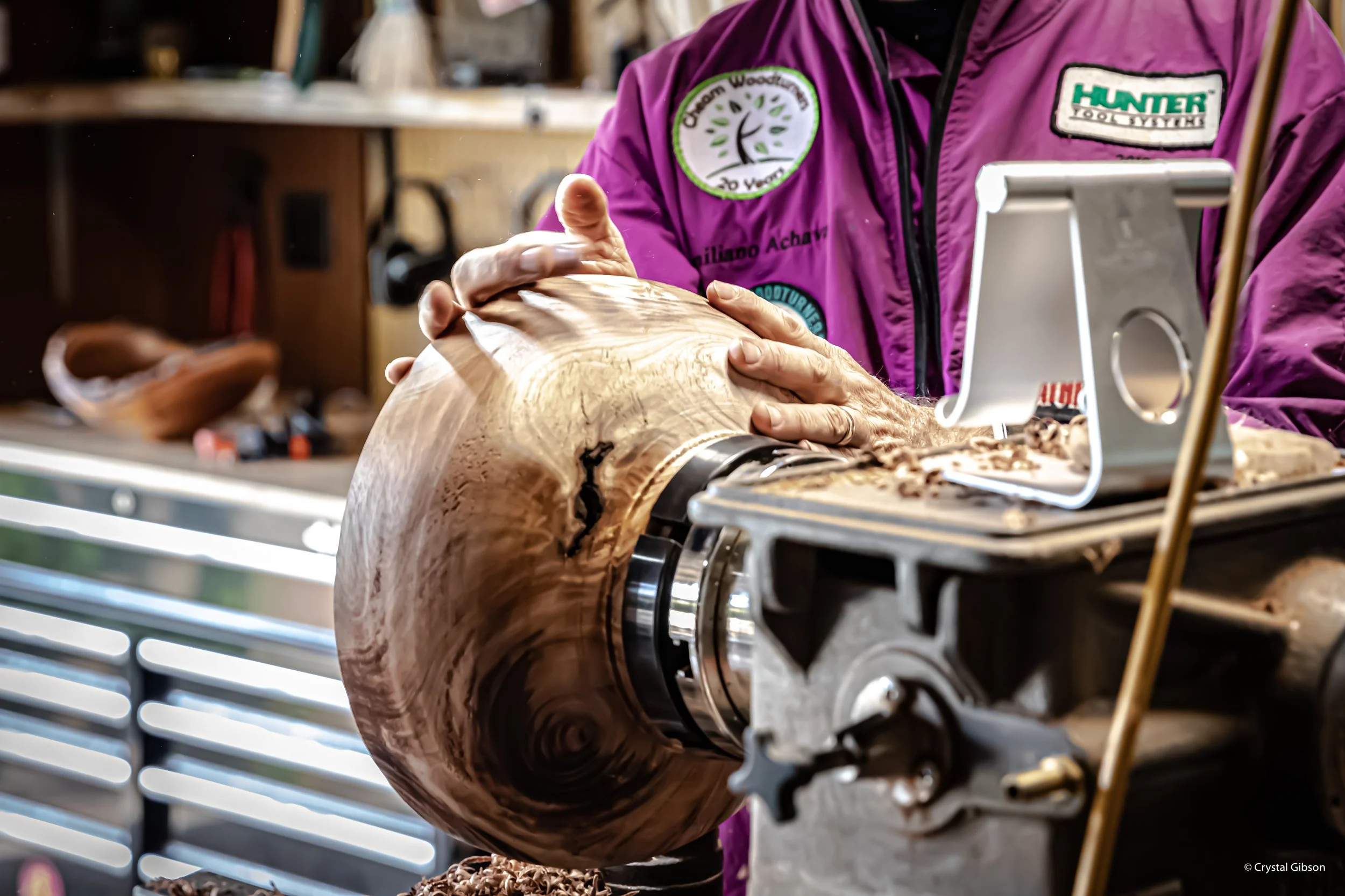 A woodworker smoothing the surface of a large, curved wooden bowl on a lathe machine.