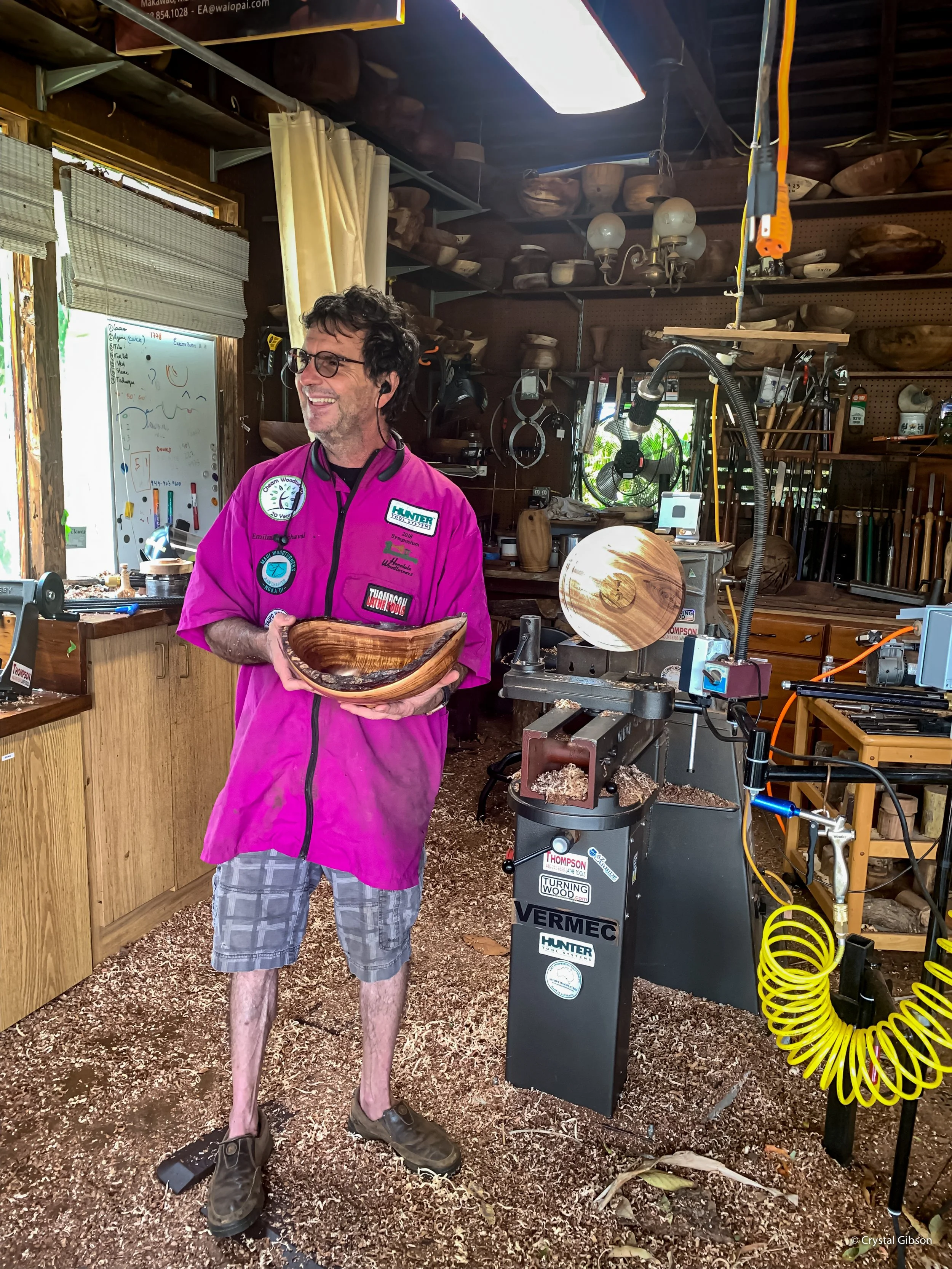 A man in a pink apron is holding a wooden bowl, standing in a woodworking shop with wood shavings on the floor and various woodworking tools and equipment surrounding him.