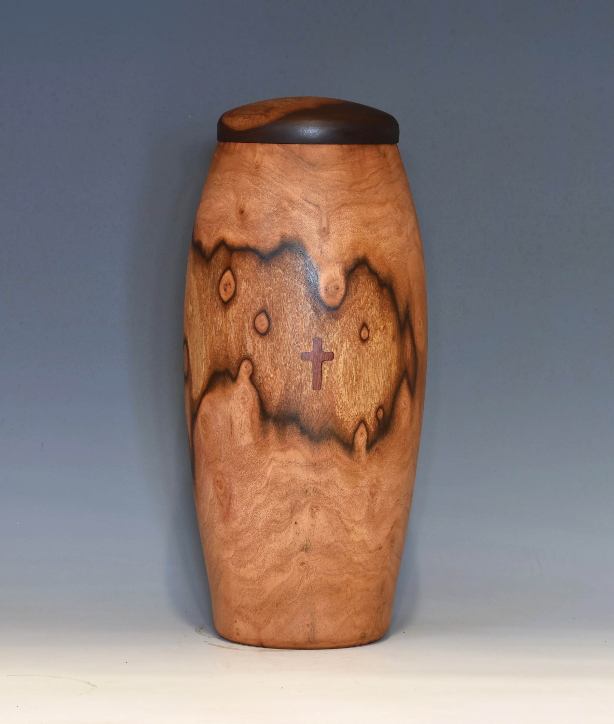 Wooden urn with a cross symbol on the front, smooth finish, and a dark lid, placed against a plain grey background.