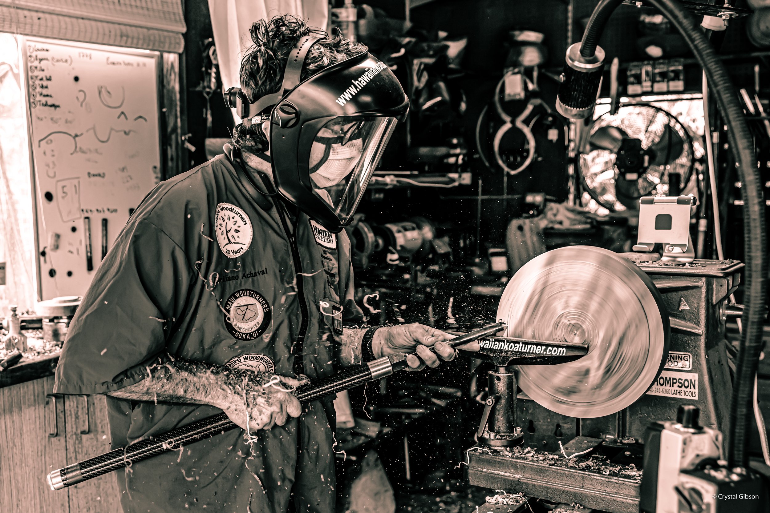 A person wearing a face shield, face mask, and work jacket is using a lathe machine to cut a piece of wood or metal. The work space is filled with equipment and tools.