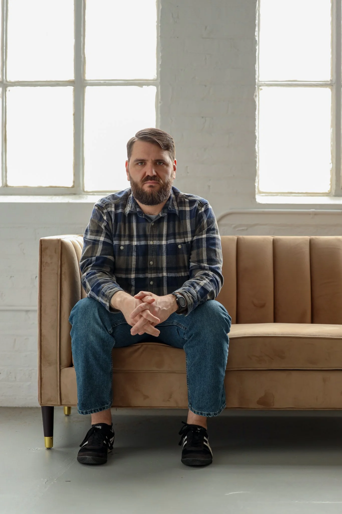 A white man with short brown hair and a short brown beard sits on a tan couch, which is front of a brick wall painted white. Sunlight is coming through large windows behind him. He's wearing a long-sleeve flannel shirt, blue jeans, and black shoes.