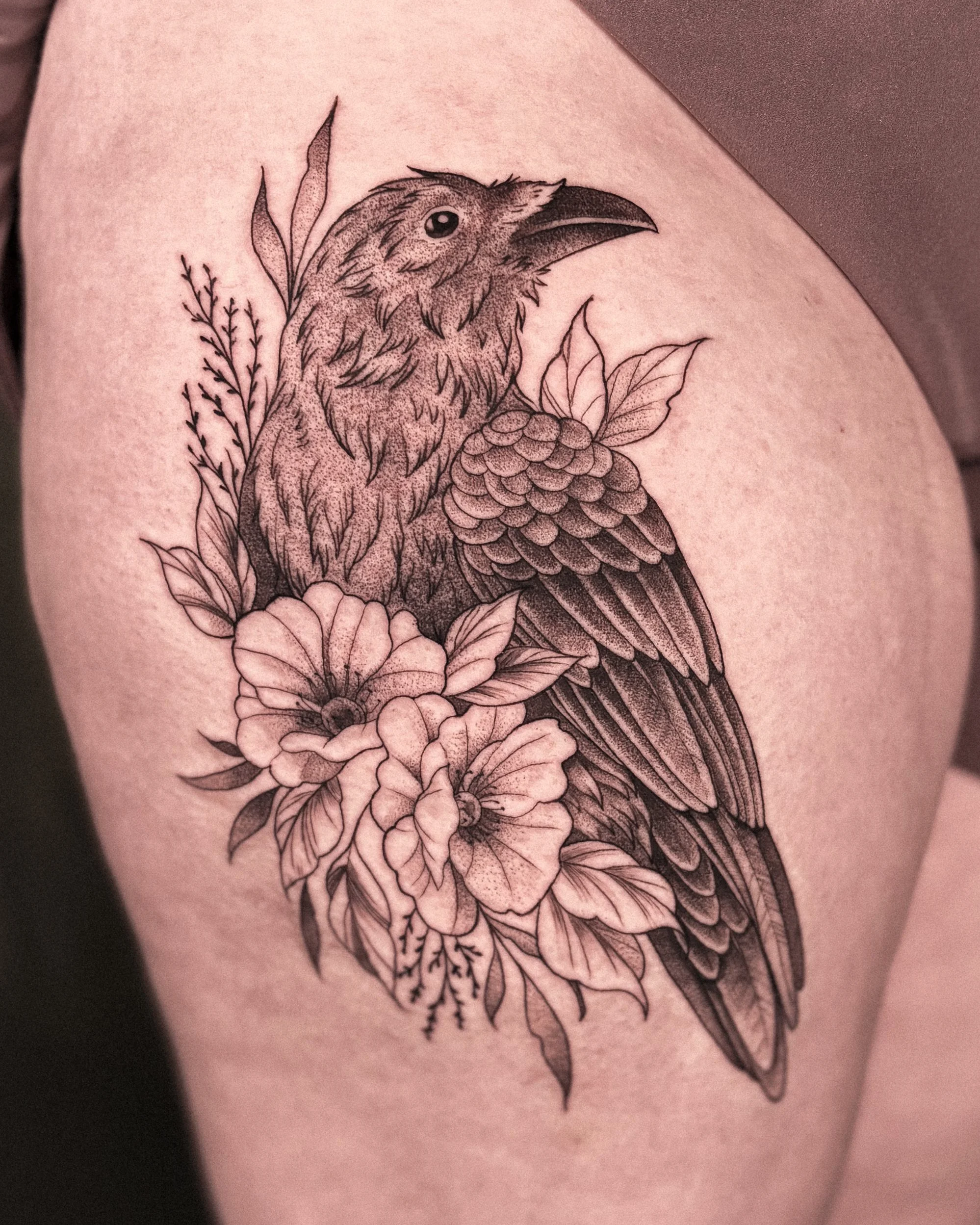 Black ink drawing of a bird, possibly a crow or raven, perched among flowers and leaves, on skin.