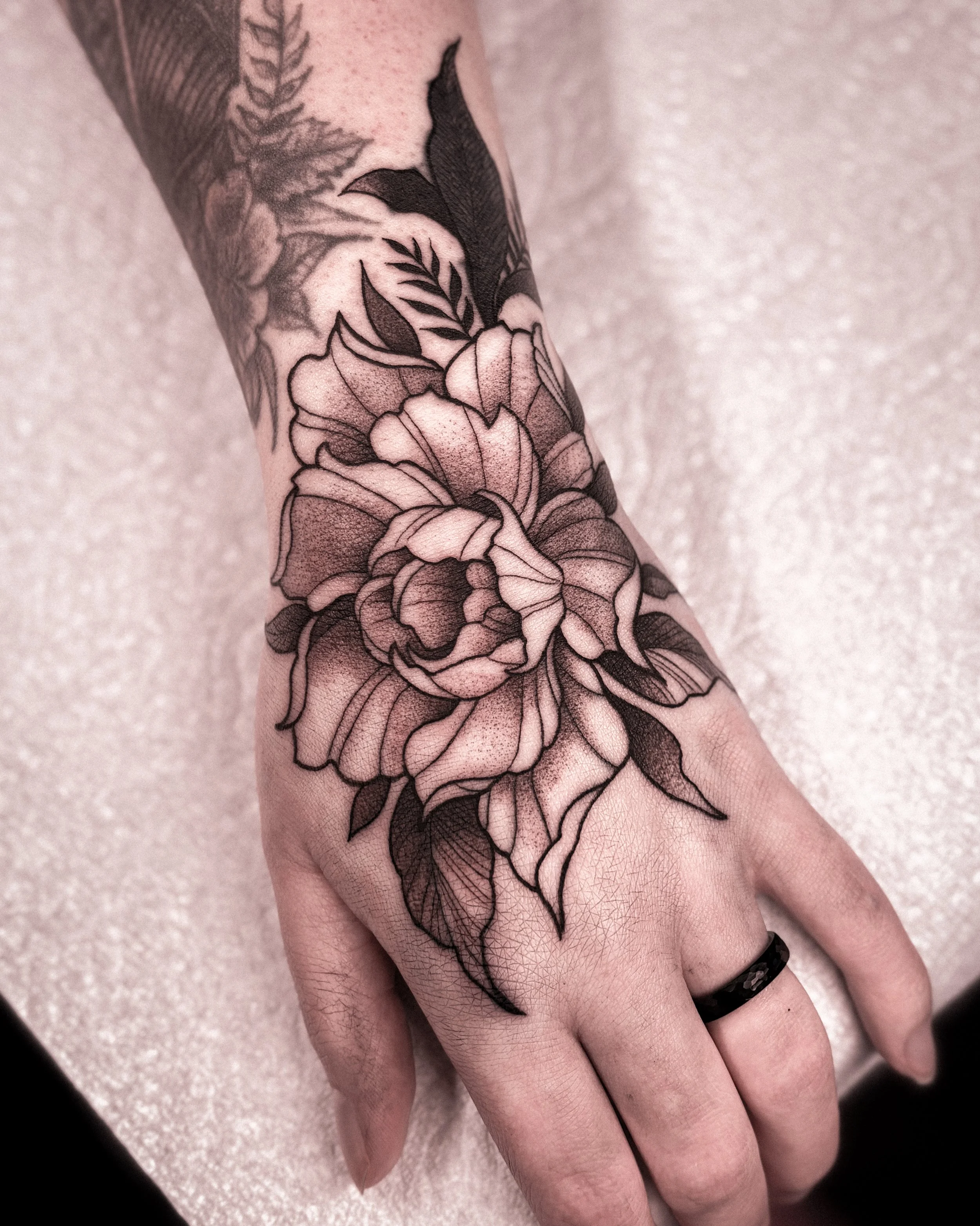 A black and gray floral tattoo on a person's hand and wrist, featuring detailed petals, leaves, and shading.