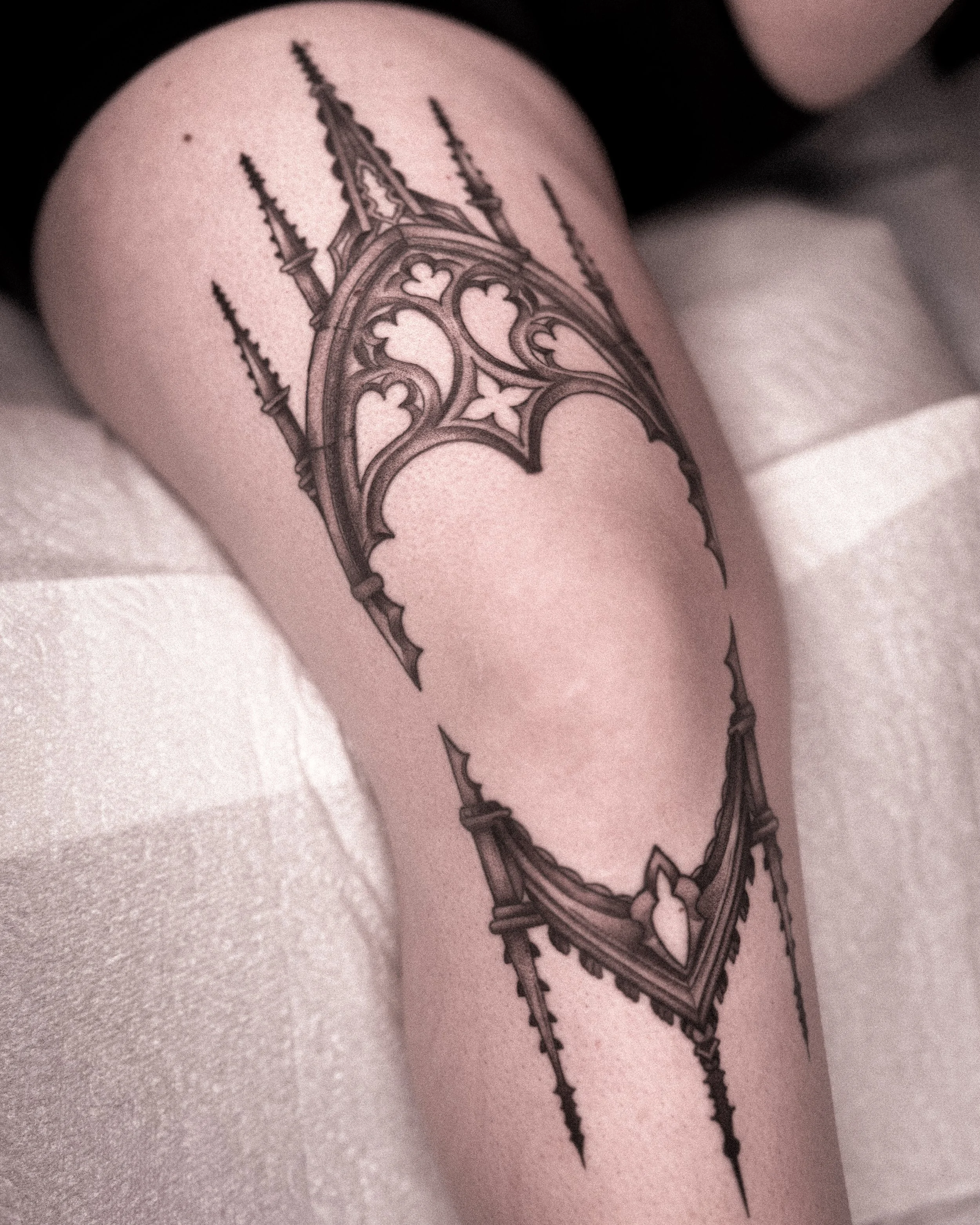 Close-up of a detailed black ink tattoo of a gothic-style cathedral and a diamond-shaped jewelry design on a person's thigh.