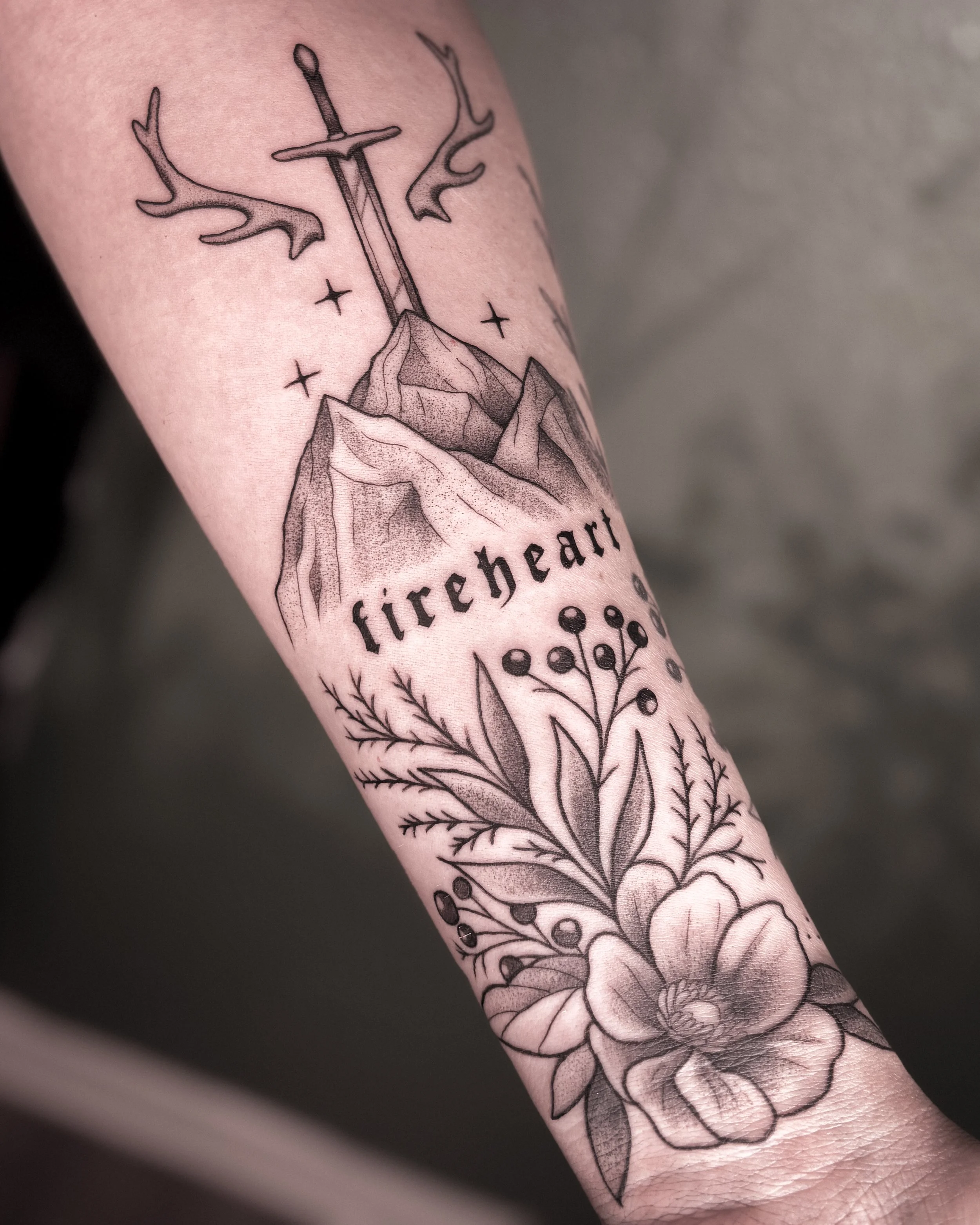 Black line tattoo of a flower with detailed petals and leaves on a person's arm, with stars and a mountain in the background.