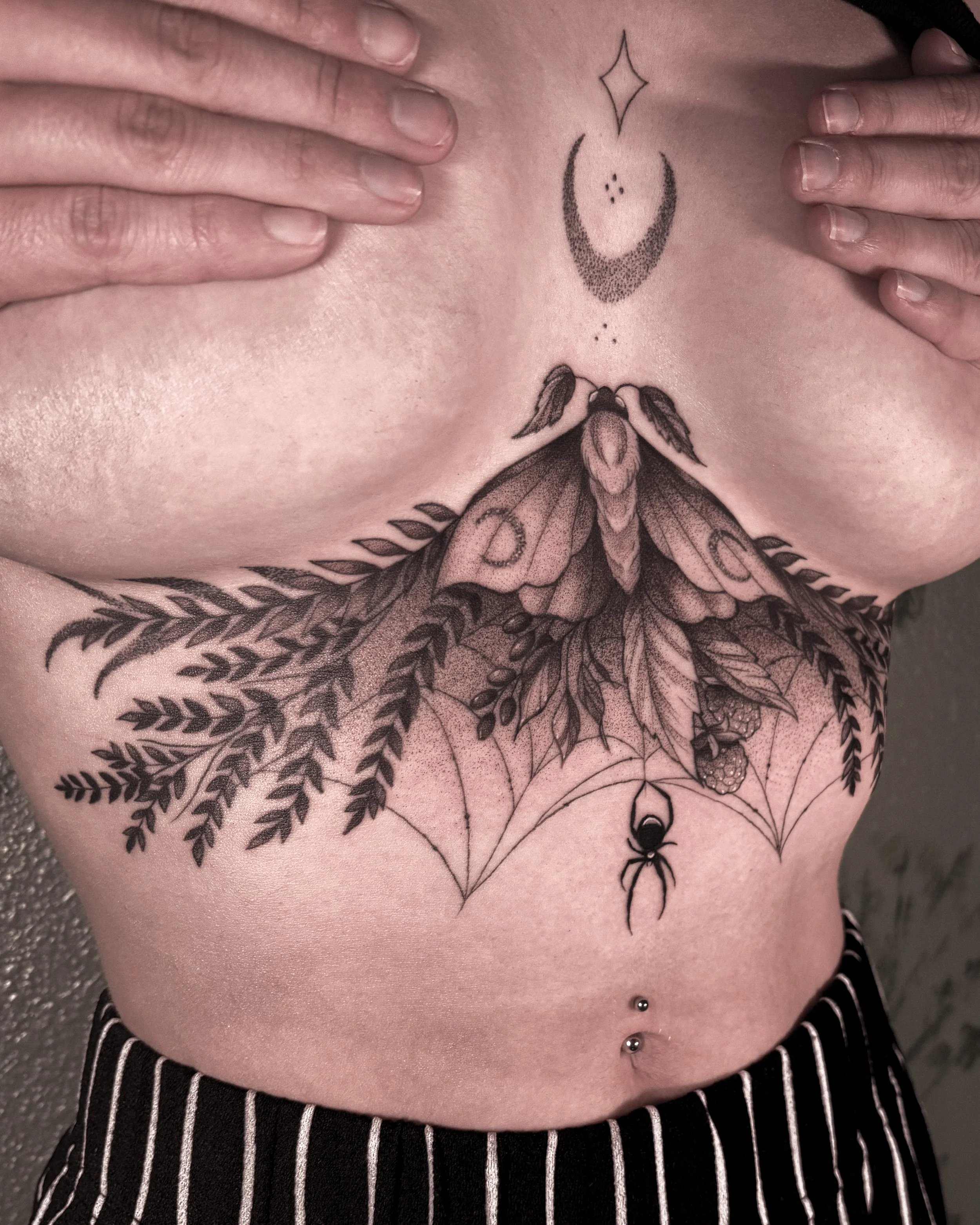 Close-up of a person's chest showing tattoos of a crescent moon with dots, a star, and a moth with outspread wings, with hands touching the chest, wearing striped clothing and jewelry.