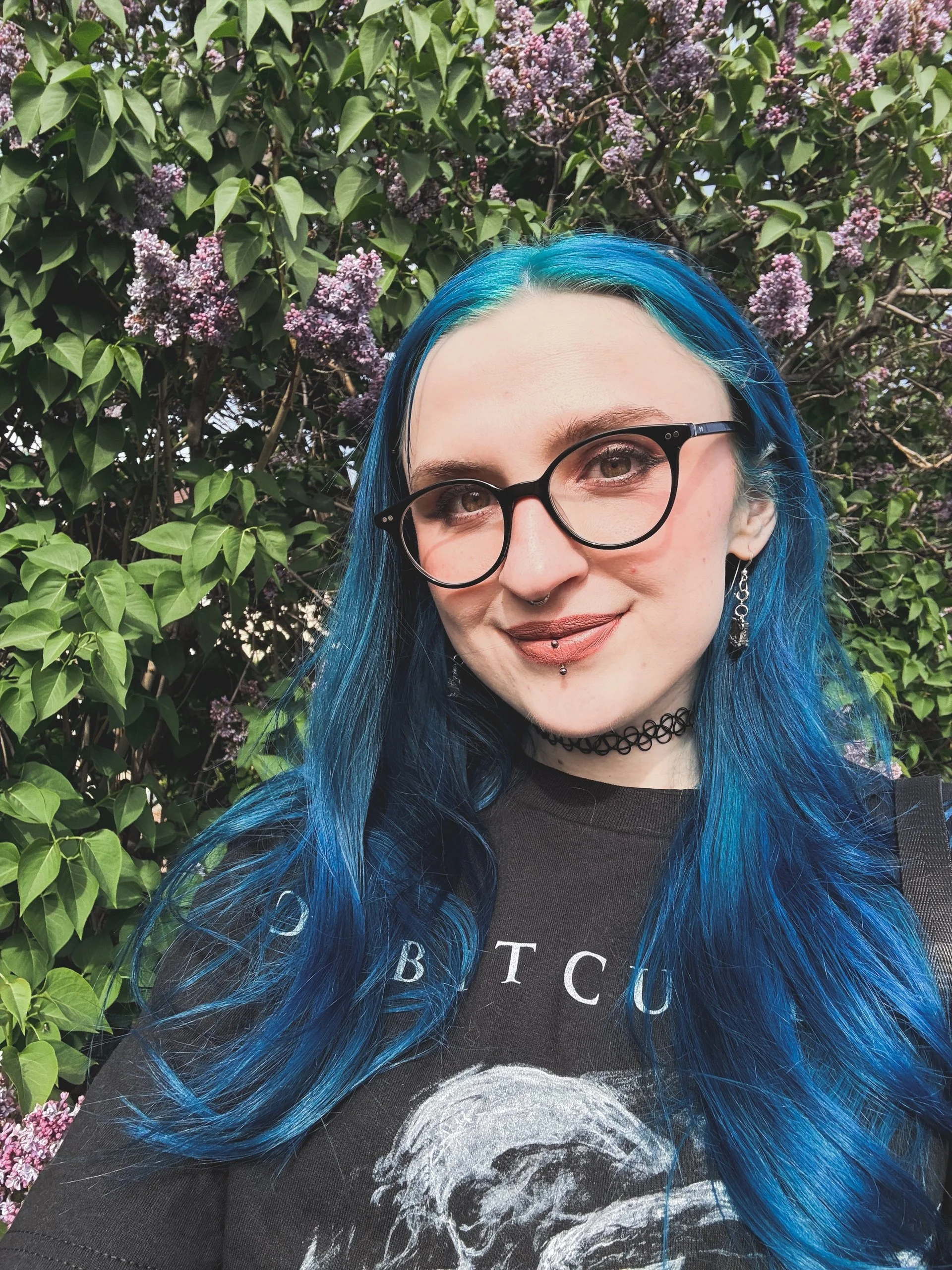 A woman with long, vibrant blue hair, black round glasses, and piercings, smiling outdoors in front of a shrub with purple flowers.