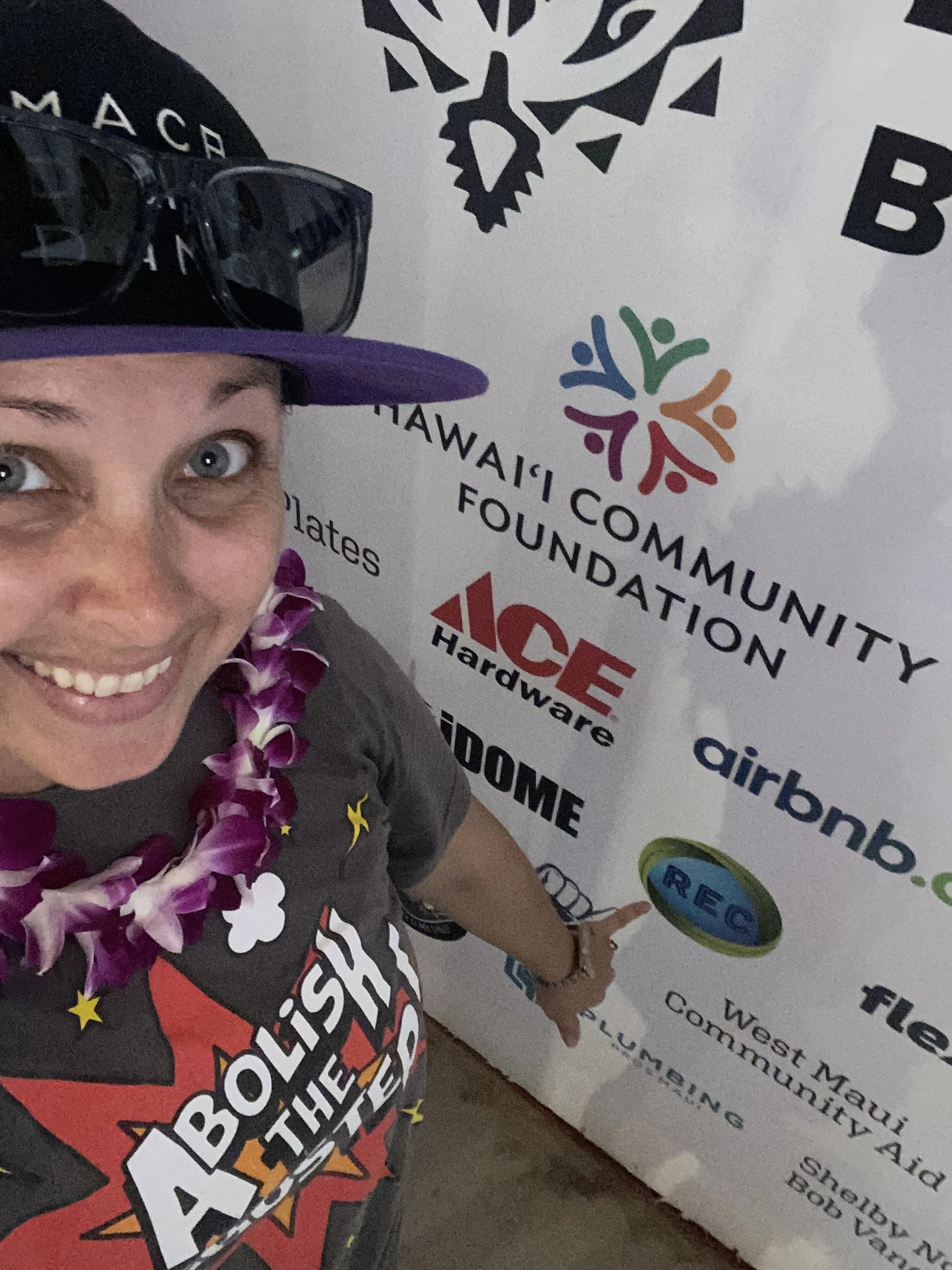 Jackie smiles at the camera wearing an orchid lei and her sunglasses placed on her hat, pointing a shaka at 'West Maui Community Aid.'