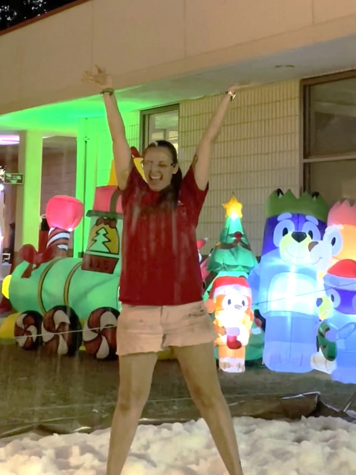 Jackie standing on "snow" in a red shirt and pink shorts in front of inflatable Christmas decorations