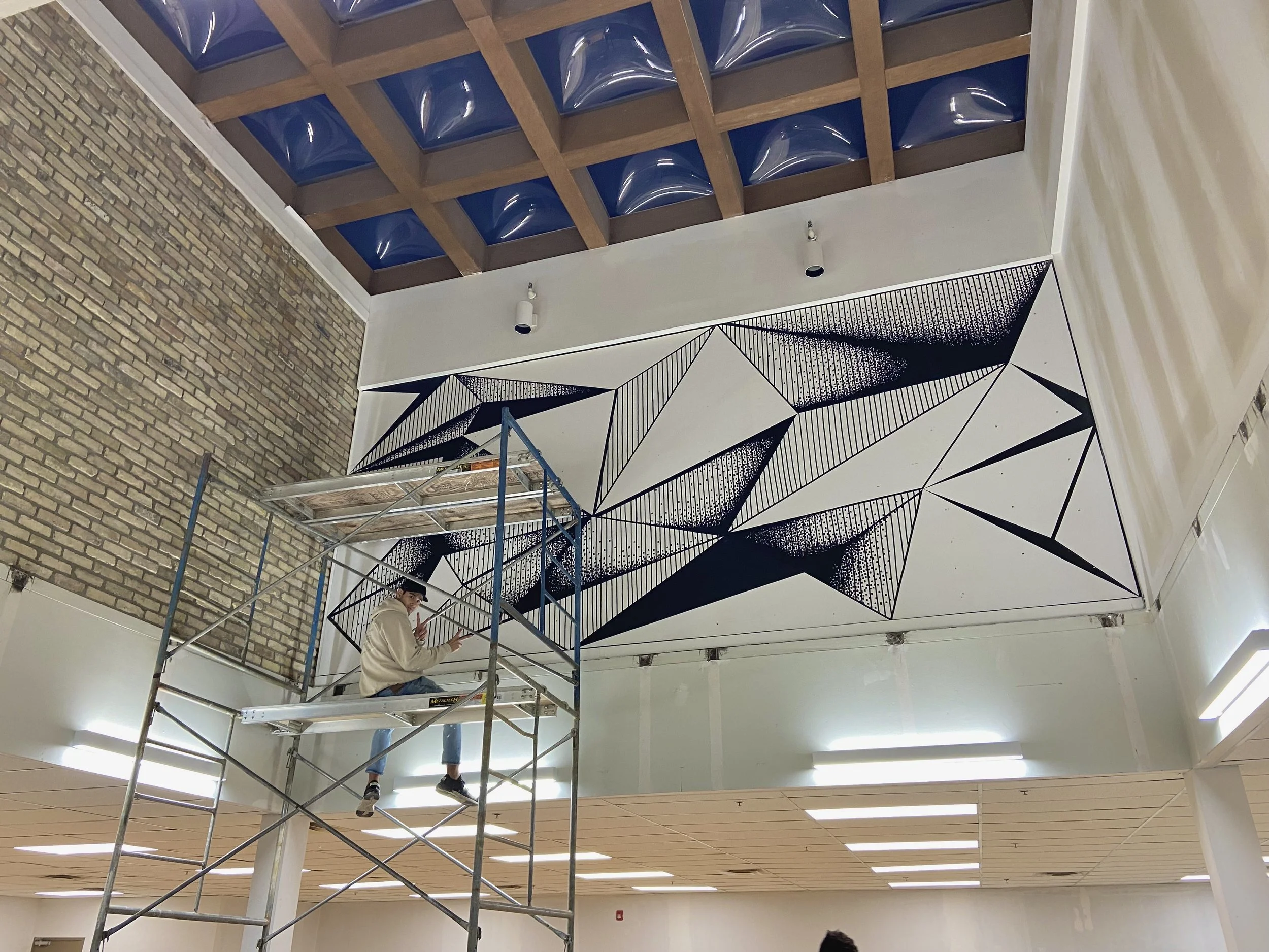 A person on scaffolding painting a large geometric mural on an indoor wall, with a brick wall on the left and a grid of blue and wooden ceiling tiles above.
