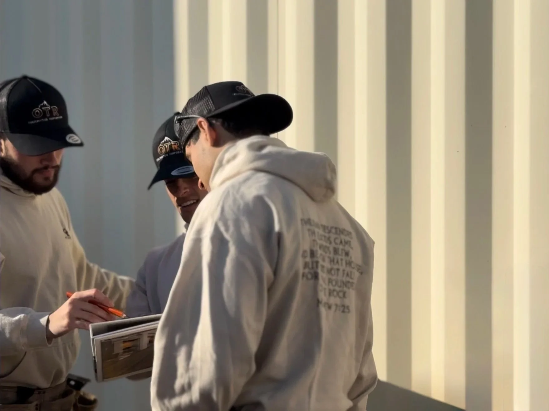 Three men wearing baseball caps, hoodies and jeans are gathered together looking at a building plans on the job site.