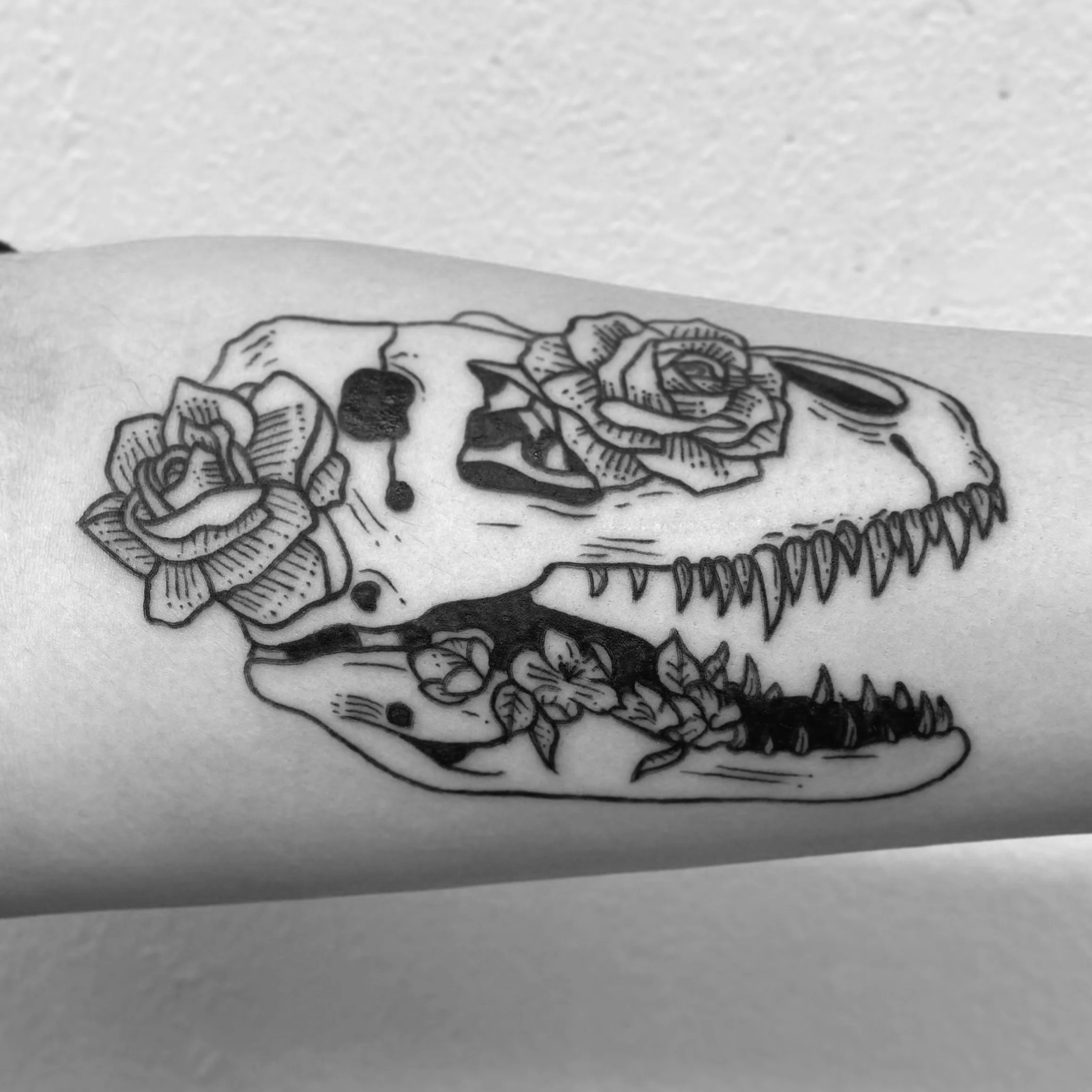 Black ink tattoo of a T. Rex skull and flowers.