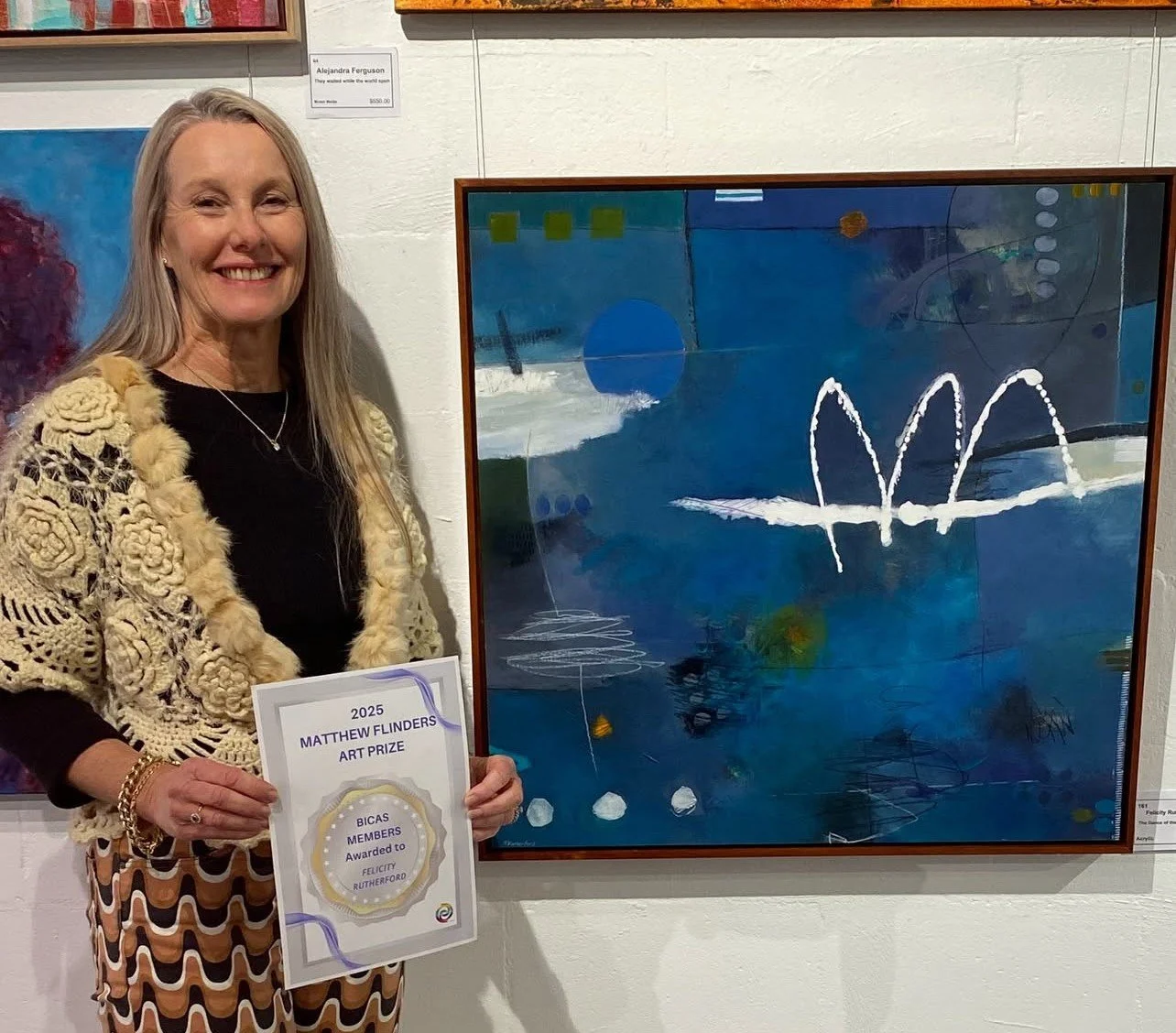 Felicity holding an art award standing next to an abstract painting on display at an art gallery.