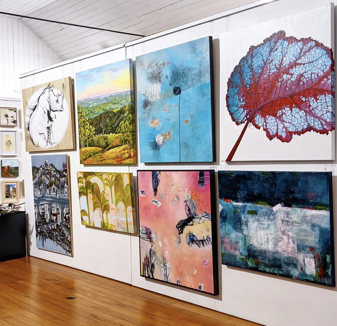 Various colorful paintings displayed on a white gallery wall, including landscape scenes and abstract art.
