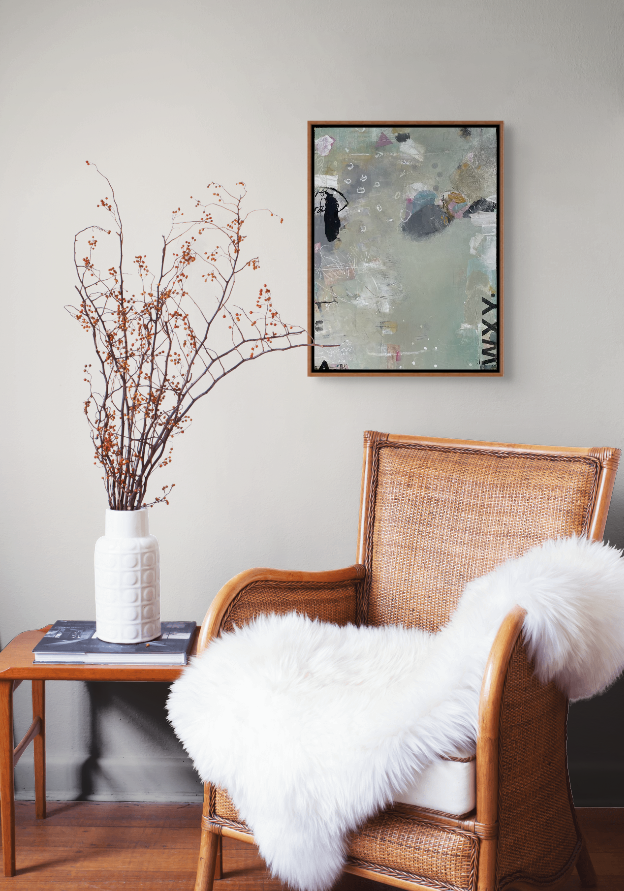 A cozy reading nook with a wicker armchair draped with a white fluffy throw, a side table holding a white vase with dried branches, and an abstract painting on a gray wall.