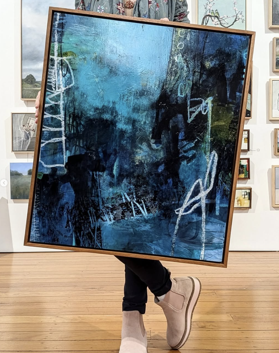 Person holding a large, framed abstract painting with shades of blue, black, and white, in an art gallery.