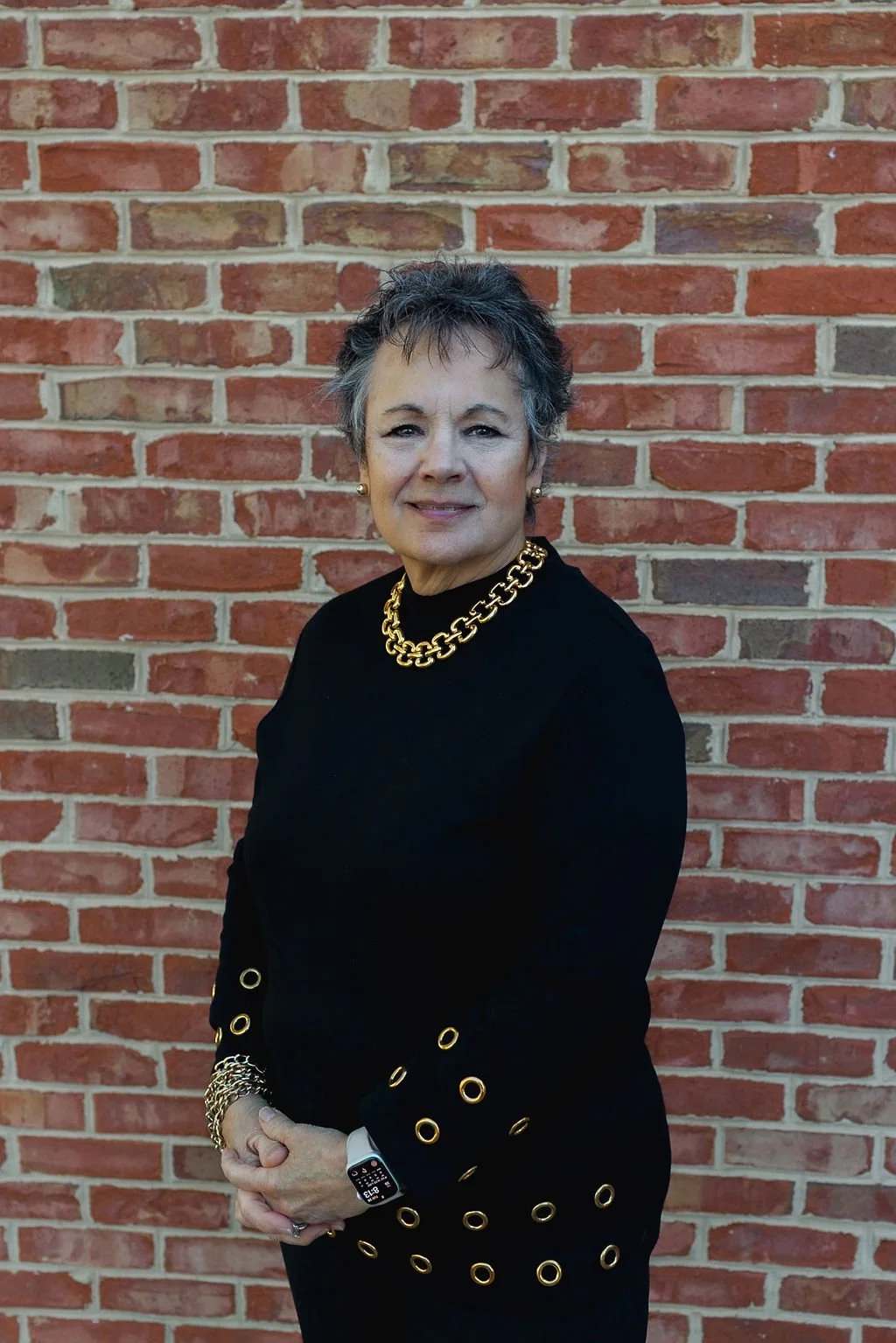 A woman with short, gray hair and earrings, wearing a black top with gold eyelet details on the sleeves, a gold chain necklace, and a smartwatch, standing in front of a brick wall.