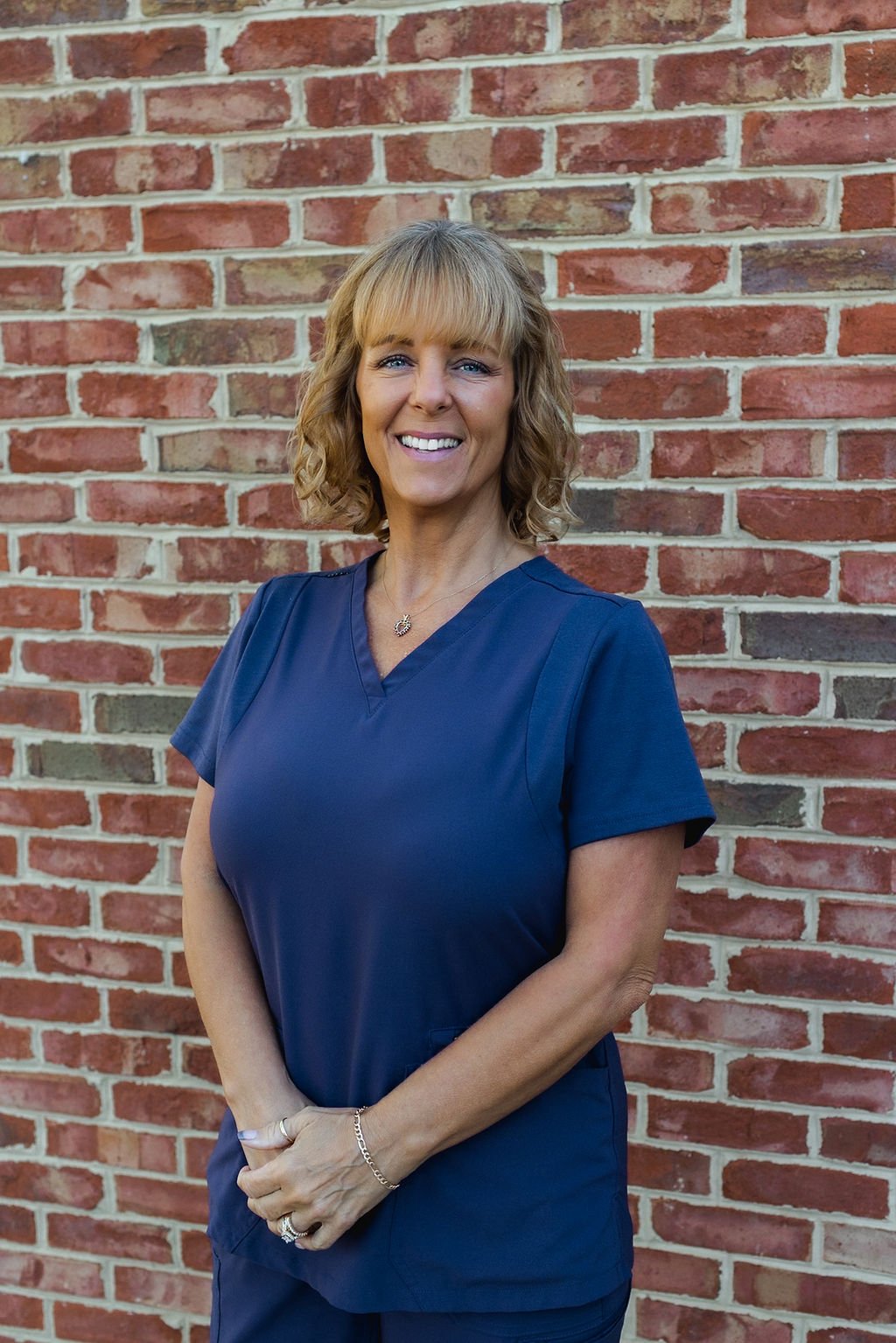 A woman wearing blue scrubs stands smiling in front of a red brick wall.