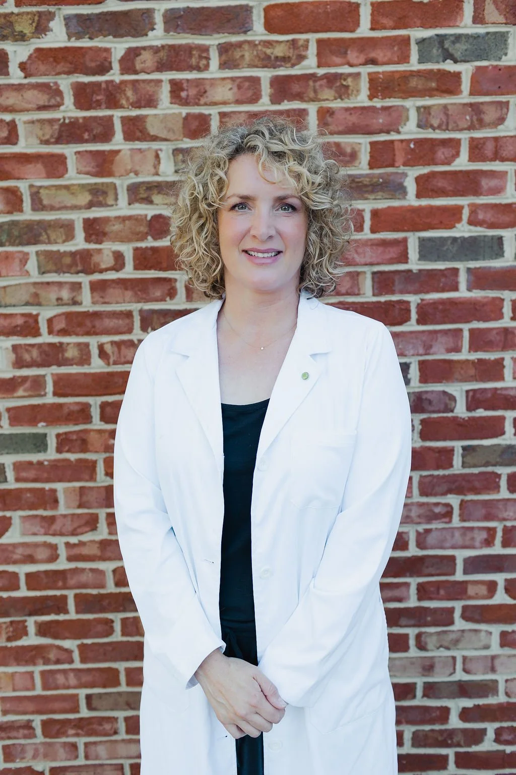 A woman with curly blonde hair wearing a white lab coat standing against a brick wall.