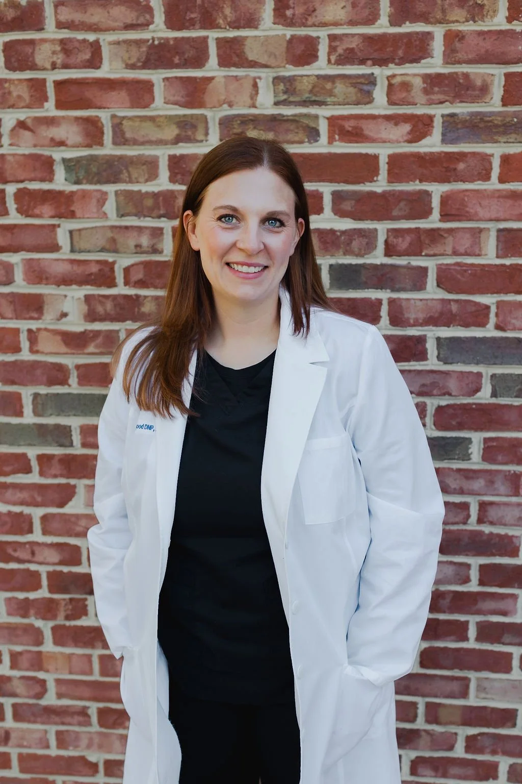 A woman wearing a white lab coat standing in front of a brick wall, smiling, with one hand in her pocket.