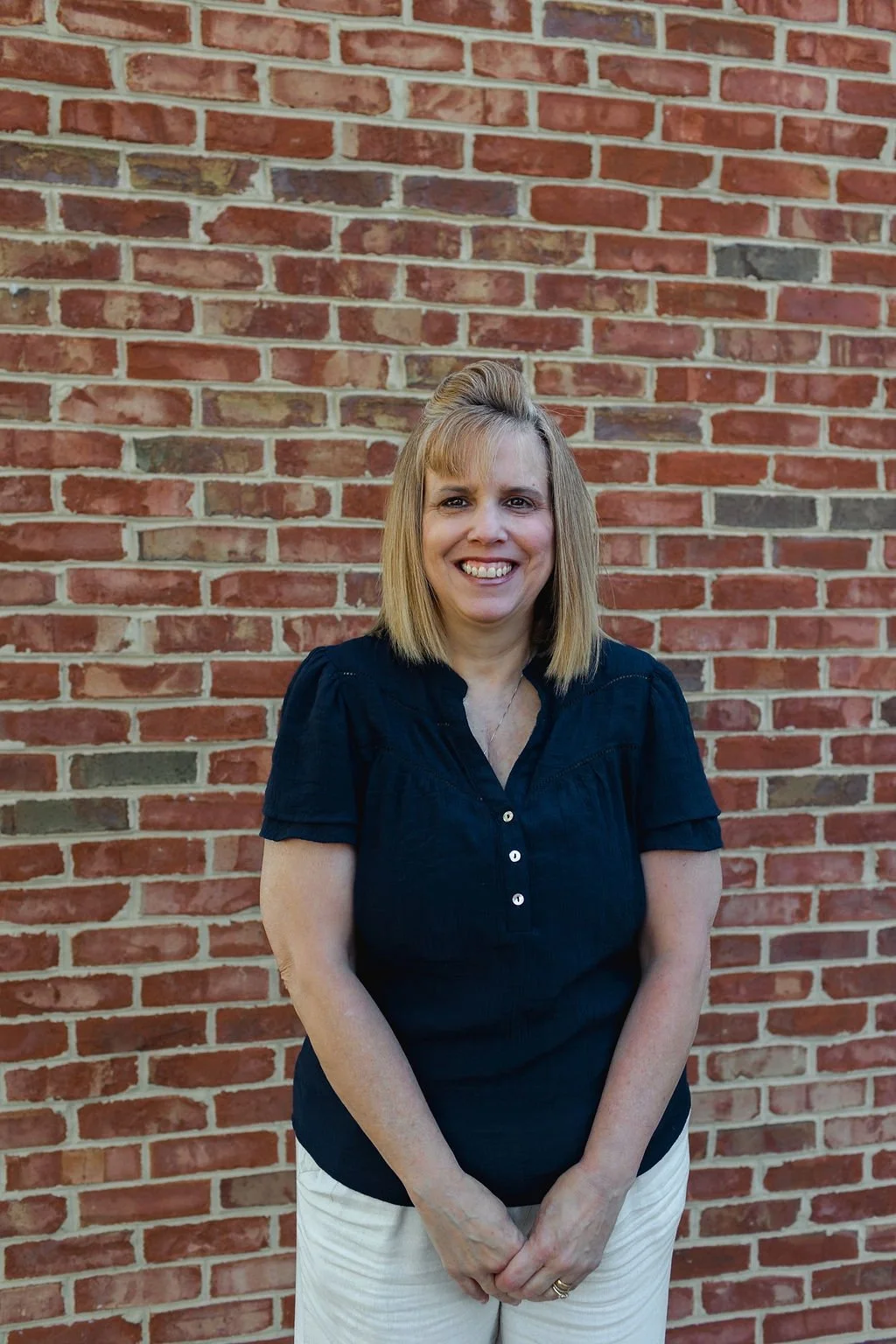 A woman with blonde hair wearing a navy blouse and beige pants standing in front of a red brick wall.