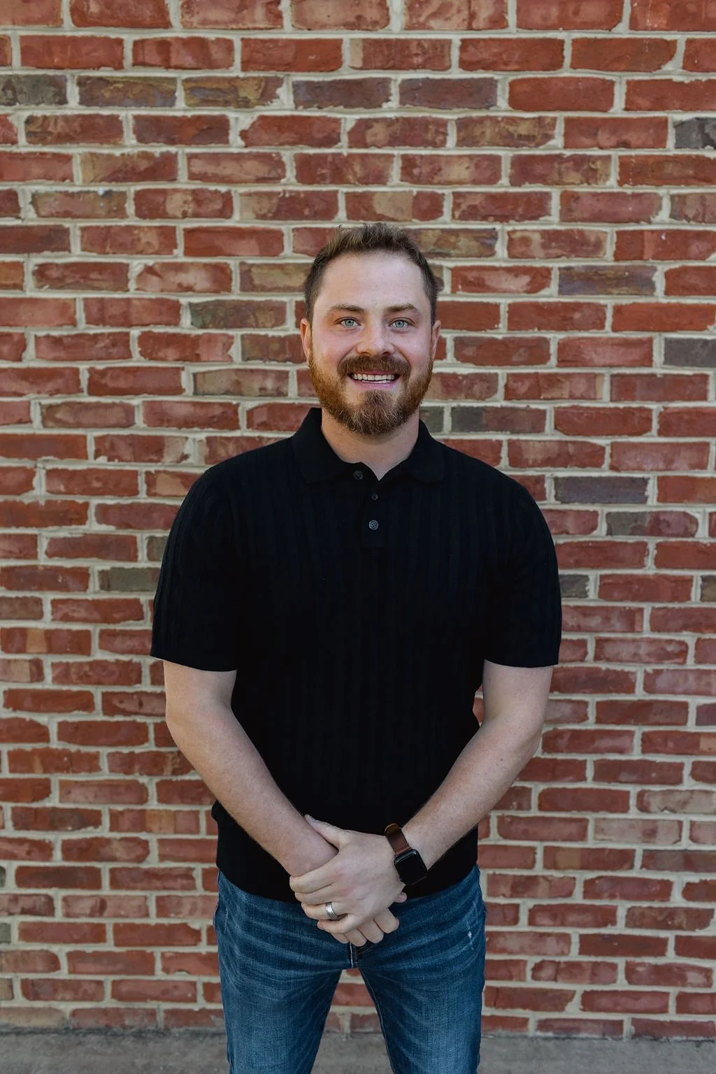 A man with light skin, a beard, and short brown hair smiling, standing in front of a brick wall, wearing a black short-sleeved polo shirt, jeans, and a black wristwatch.