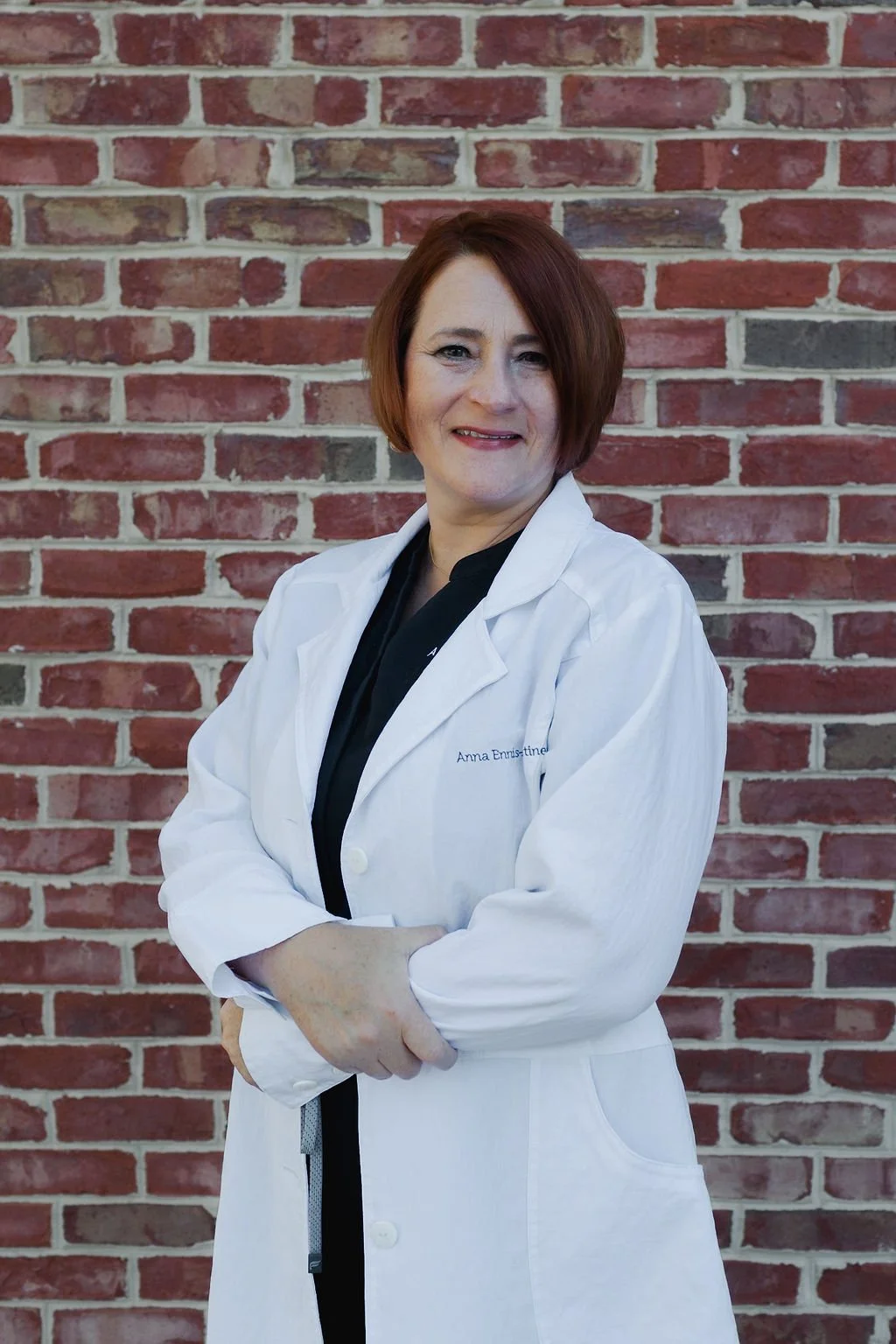 A woman with short red hair wearing a white lab coat and black shirt, standing in front of a brick wall, smiling with arms crossed.