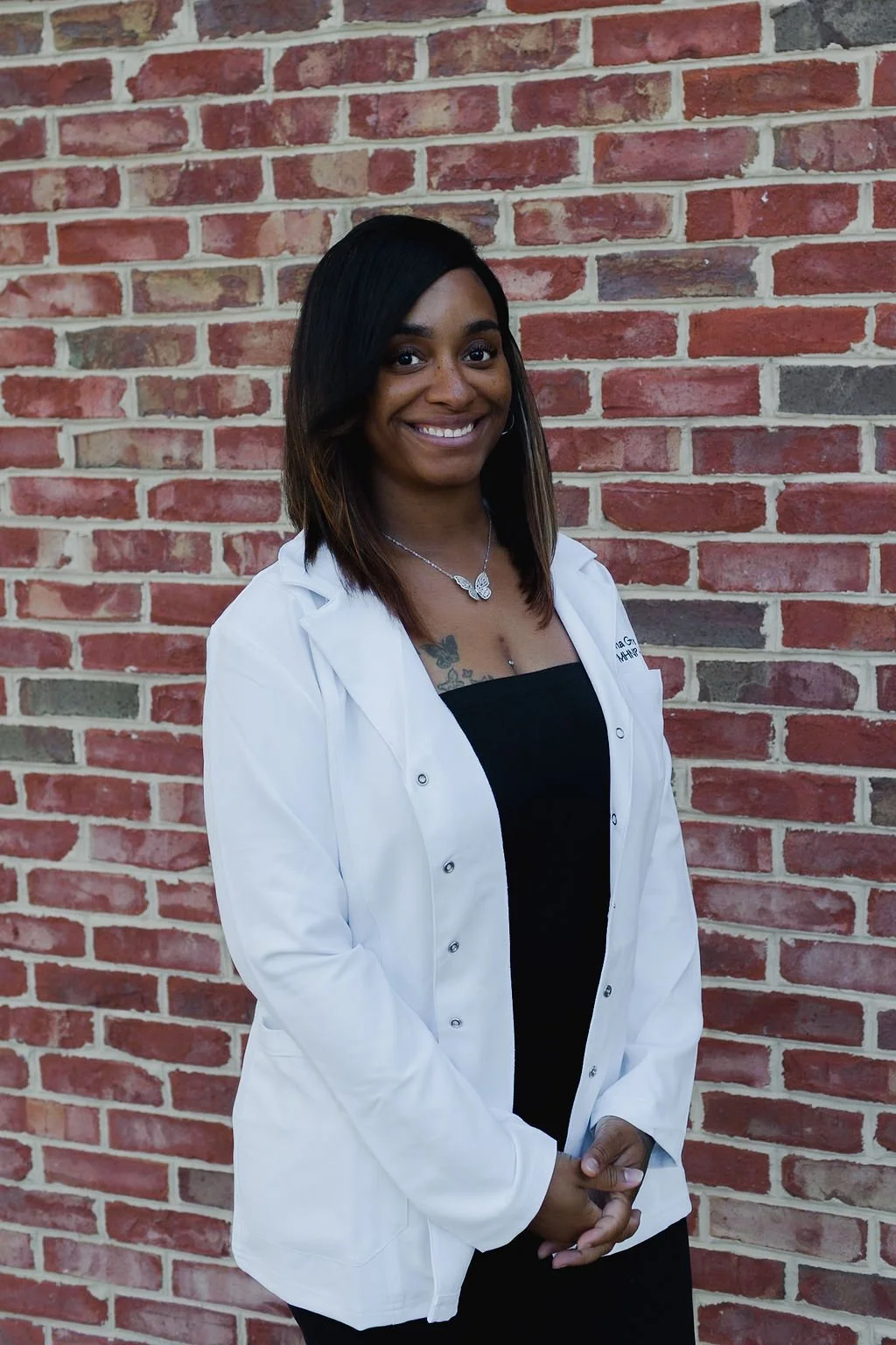 Young woman with shoulder-length black hair, wearing a white coat over a black top, standing in front of a brick wall, smiling at the camera.