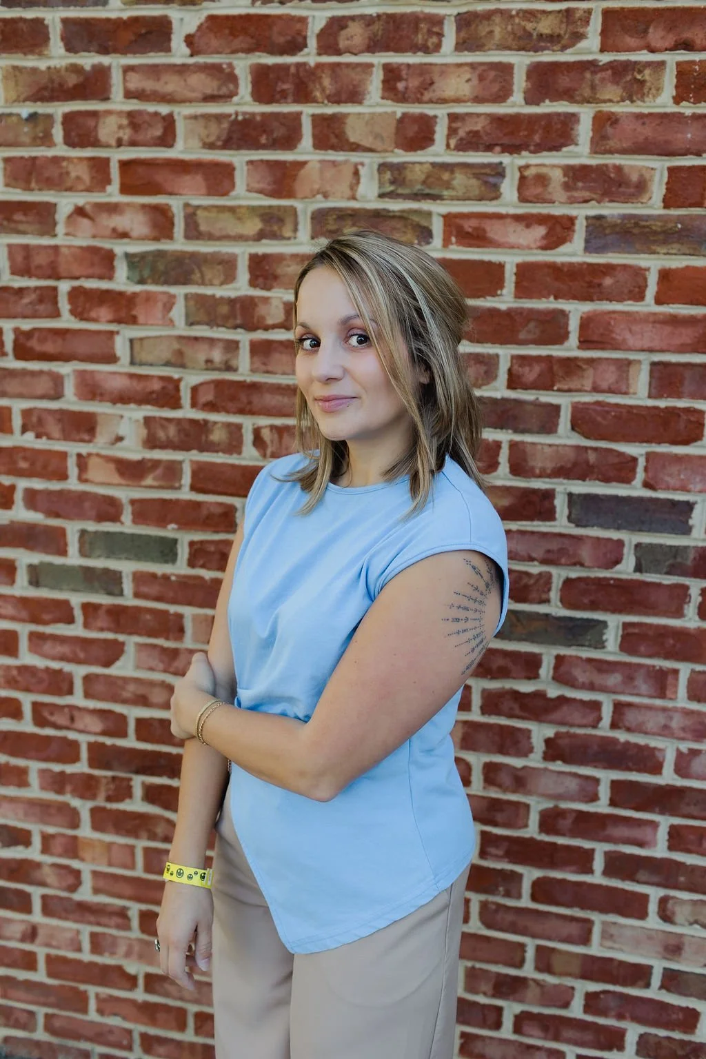A woman with shoulder-length blonde hair, wearing a light blue sleeveless top and beige pants, standing in front of a red brick wall. She has her arms crossed and is looking slightly to her left with a slight smile.