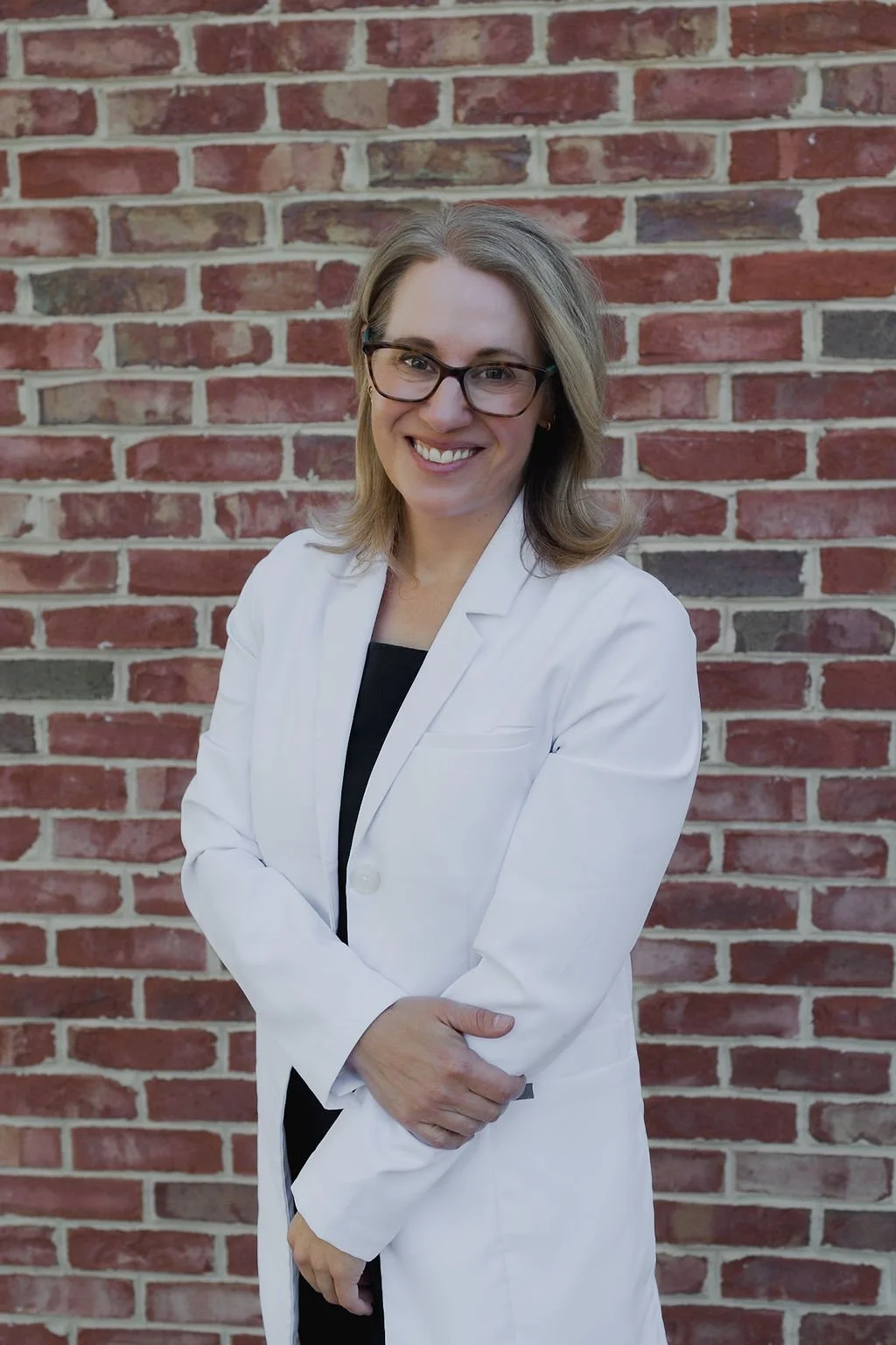 A woman with glasses and shoulder-length blonde hair wearing a white lab coat, standing in front of a brick wall and smiling.
