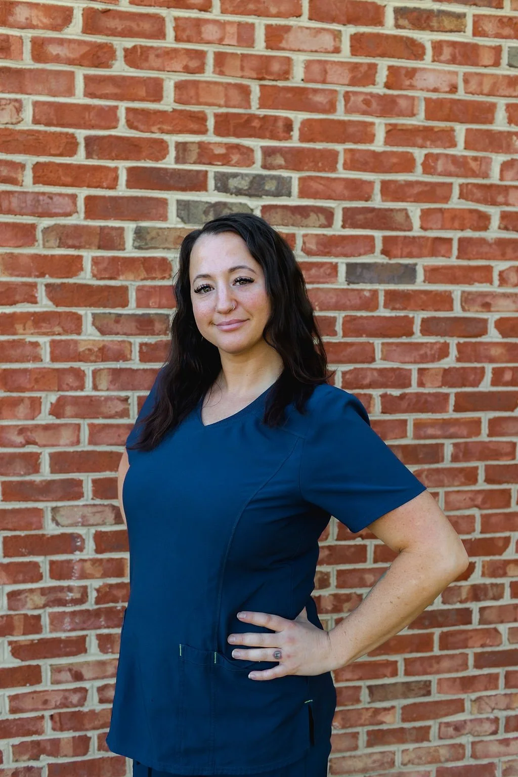 A woman with dark wavy hair, wearing a blue medical scrub top, standing with one hand on her hip in front of a brick wall, smiling slightly.