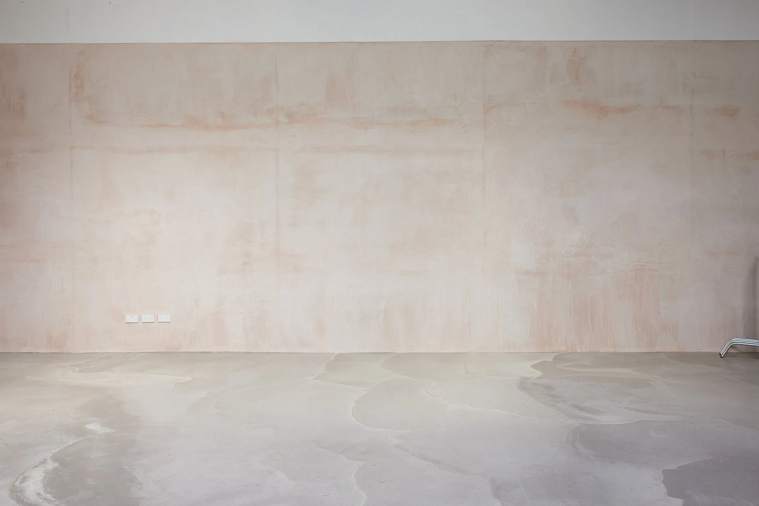 Empty room with a plain pink wall, a beige concrete floor, and electrical outlets on the lower left side.