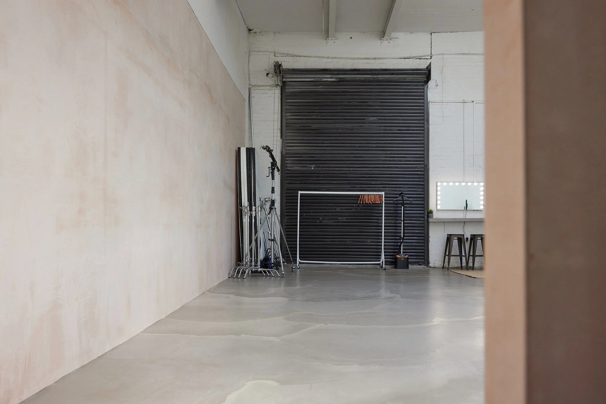 An empty photography studio or art space with a large black roll-up door, tripod, clothing rack, mirror with lights, small plant, and chairs.