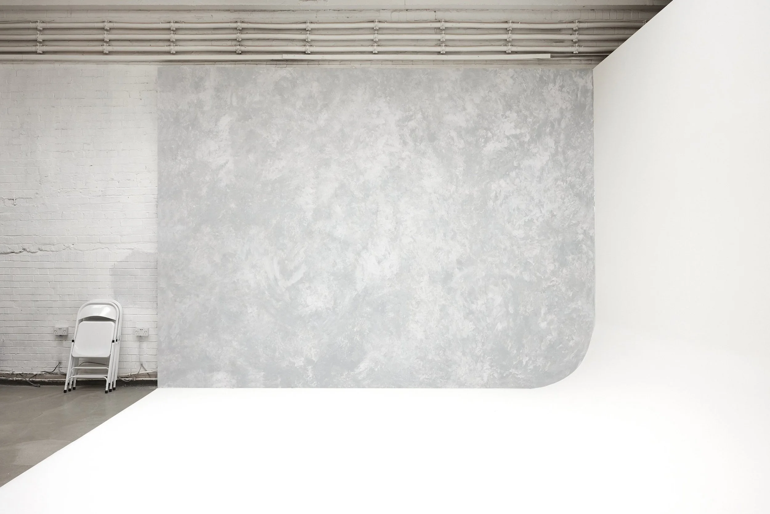 Photography studio Glasgow - Empty white and gray studio with a white floor, textured gray wall, white brick wall, and stacked white folding chairs in the corner.