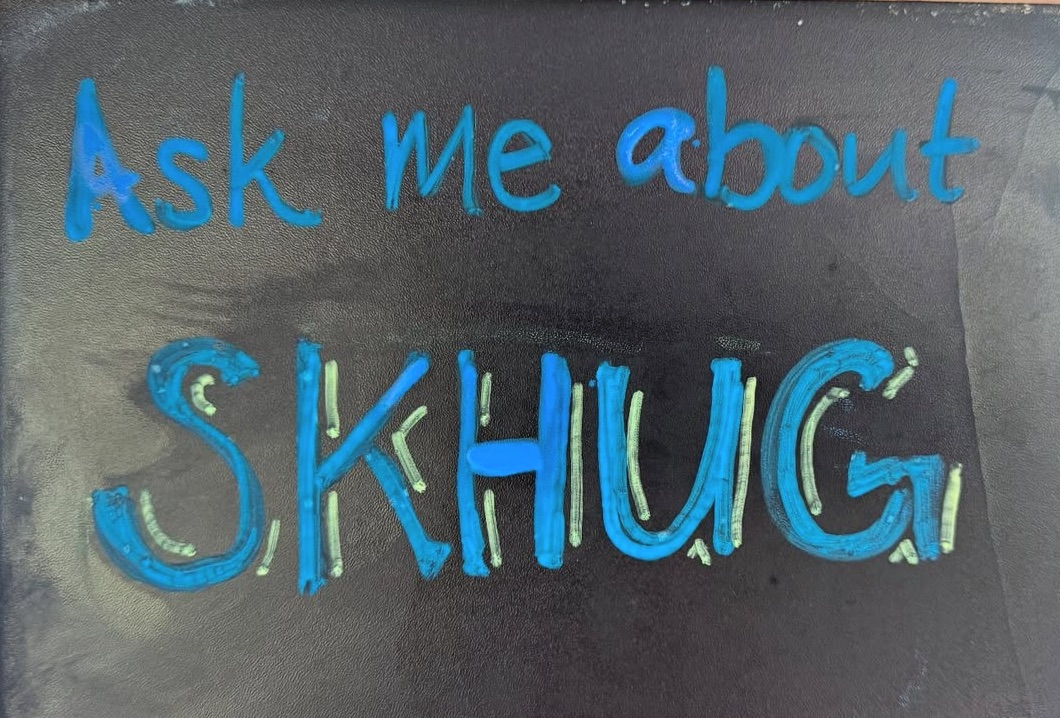 A blackboard with the message "Ask me about SKHUG!" written in blue and yellow marker.